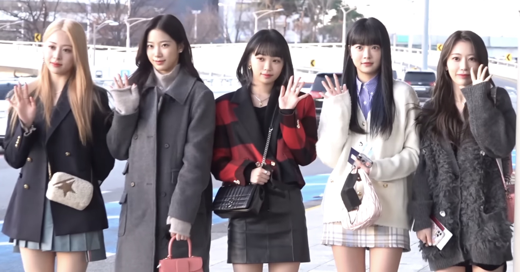 LE SSERAFIM waving at an airport departure press call in coordinated travel looks.