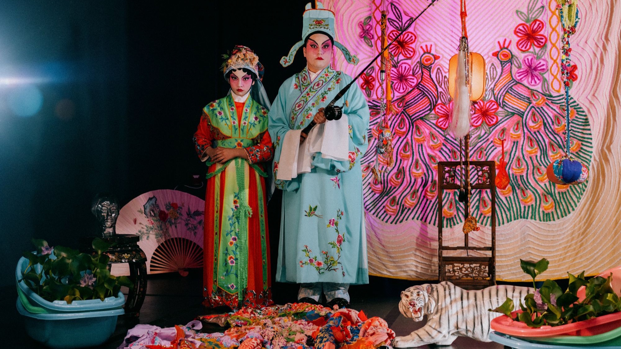 Alison Kuo, right, in a pale blue general's robe and costume beard, stands with Mei Brunette, left, in red and green opera costume, in front of a pink peacock backdrop.
