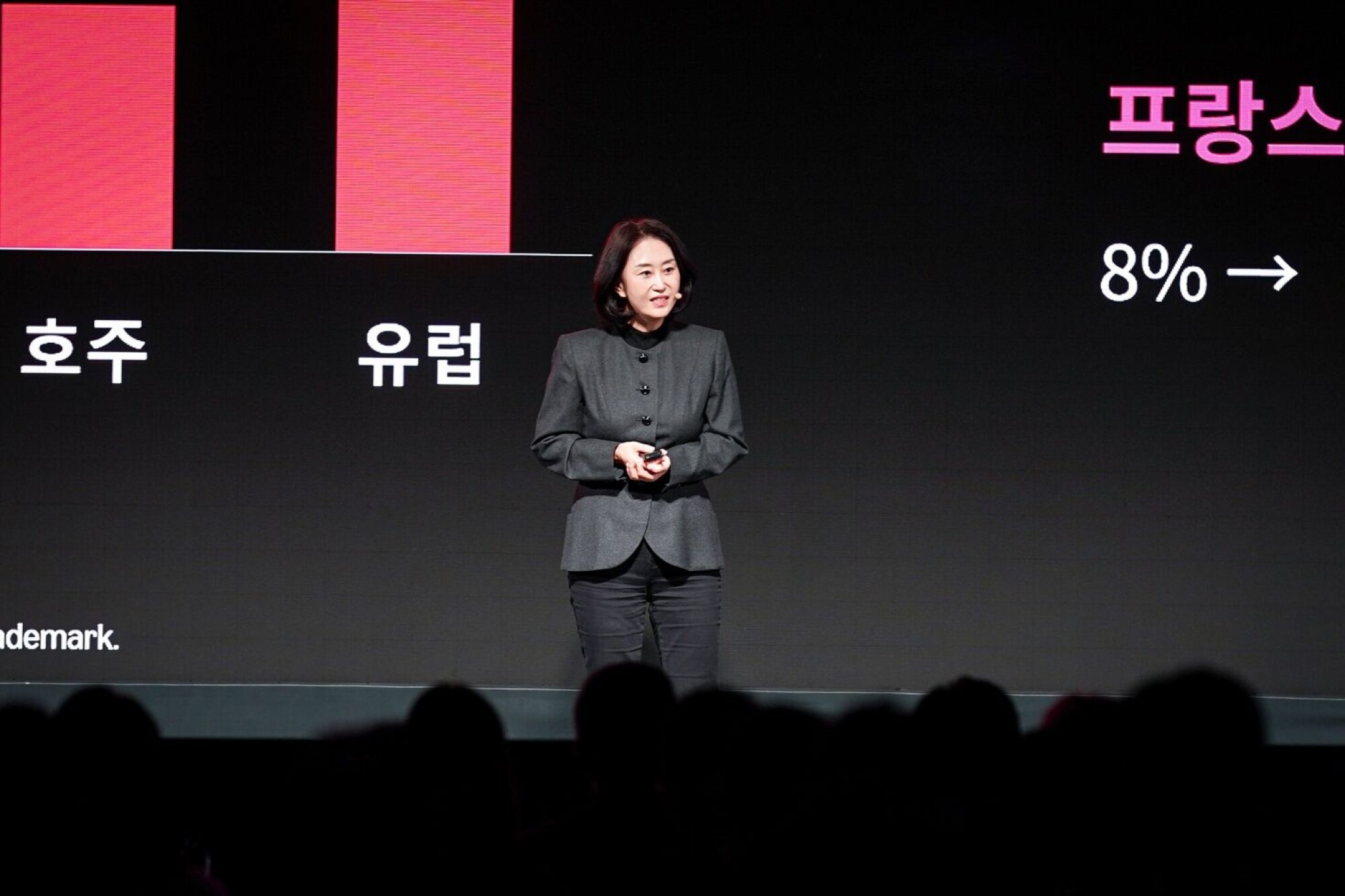 Shin Hwa-sook stands on a conference stage in a dark jacket, in front of a screen with red bars and Korean text, as an audience watches from the dark.