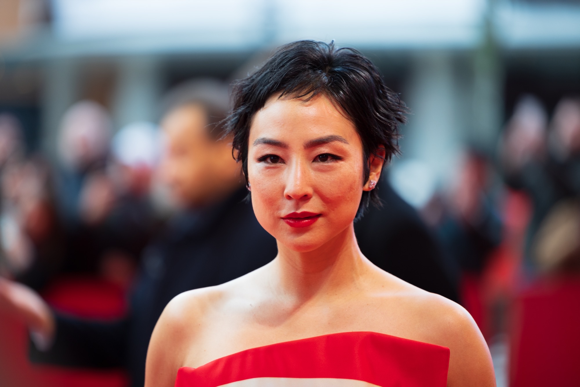 Actress Greta Lee in a red strapless gown on a festival red carpet, framed alone with a cool, intense expression against a blurred crowd.