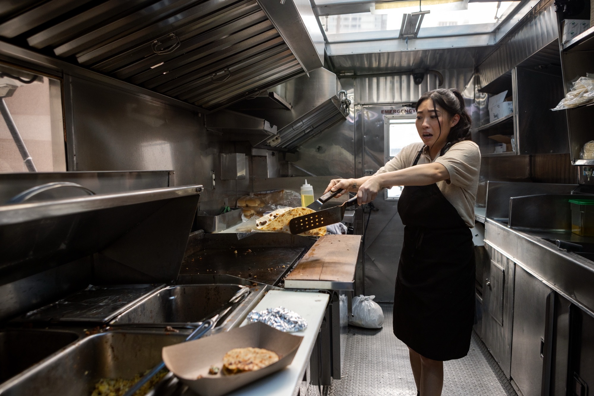 Awkwafina in a black apron flipping food on a flat-top griddle inside a stainless-steel kitchen, mid-cook with a focused expression.