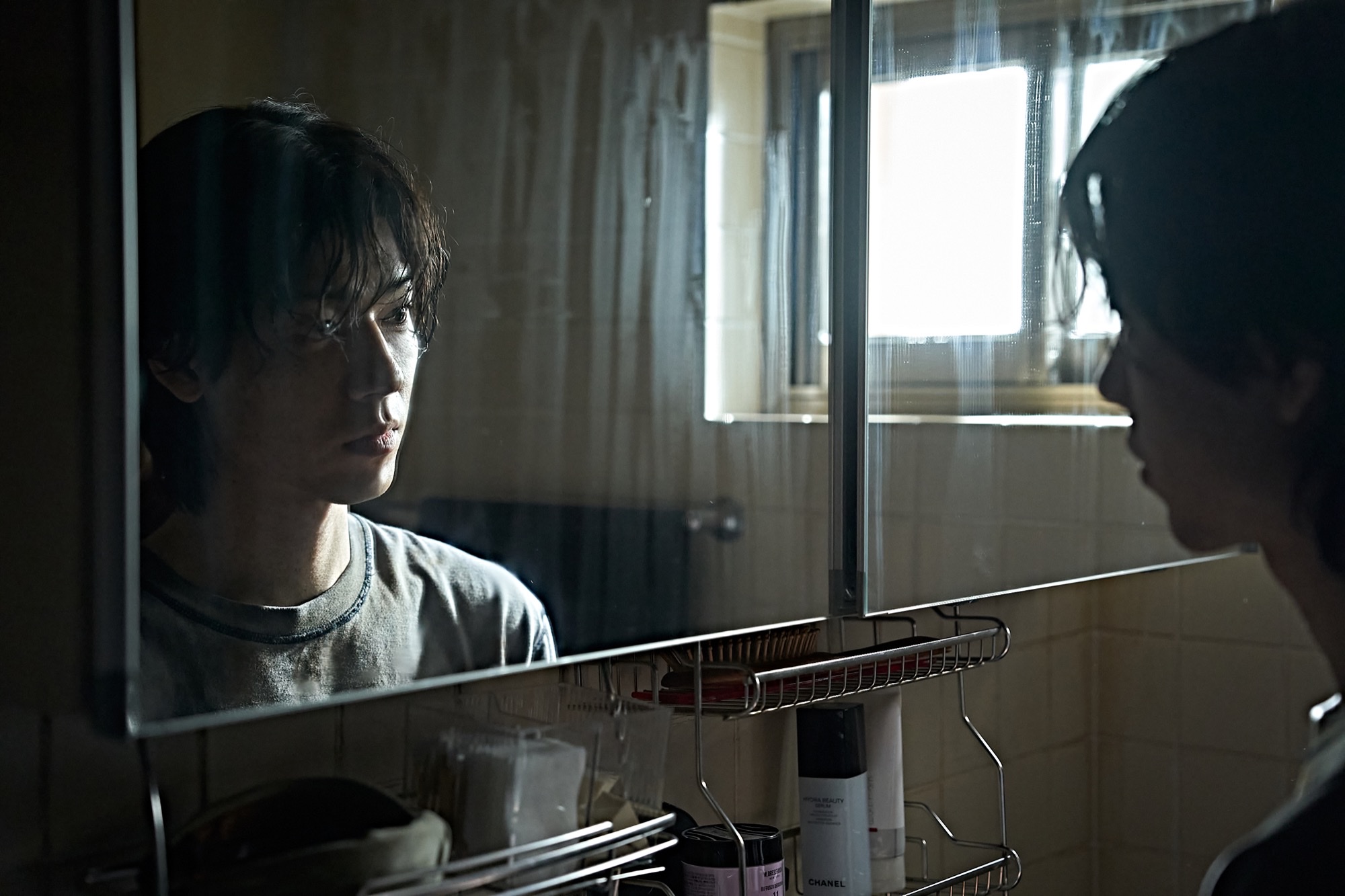 Bae Hyun-sung as Ryu Sun-woo in Paradise Lost, staring into a bathroom mirror in a grey T-shirt under hard window light.