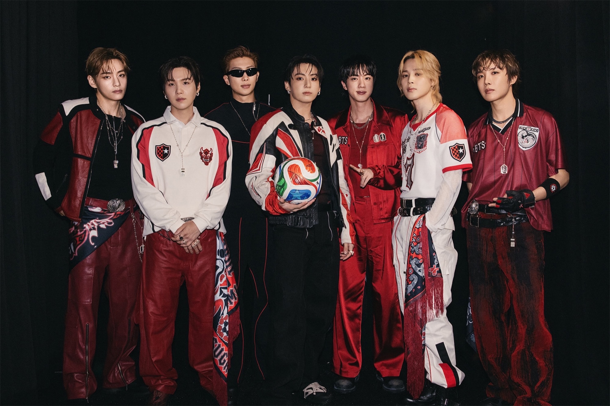BTS members pose in red, black, and white World Cup stage looks against a black backdrop, with Jungkook holding a soccer ball at center.