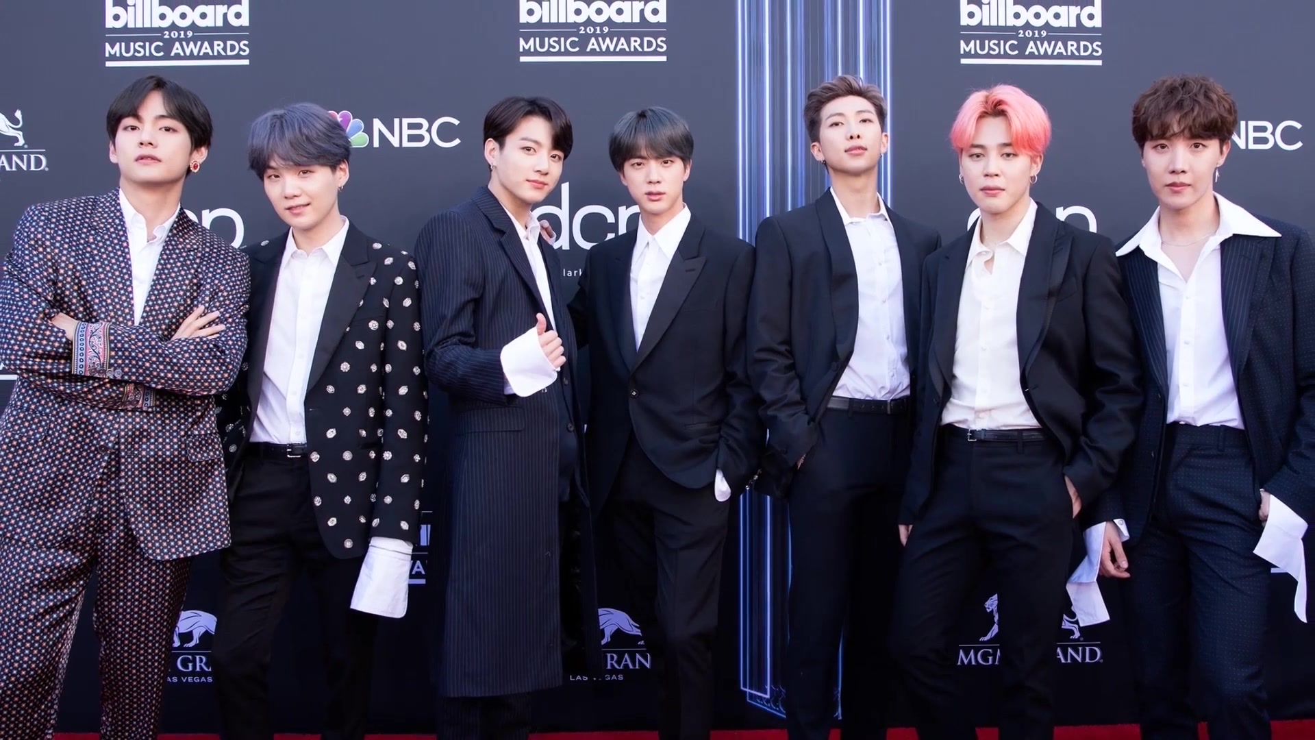 All seven BTS members in dark suits posing on the Billboard Music Awards red carpet in front of an awards-show step-and-repeat backdrop.
