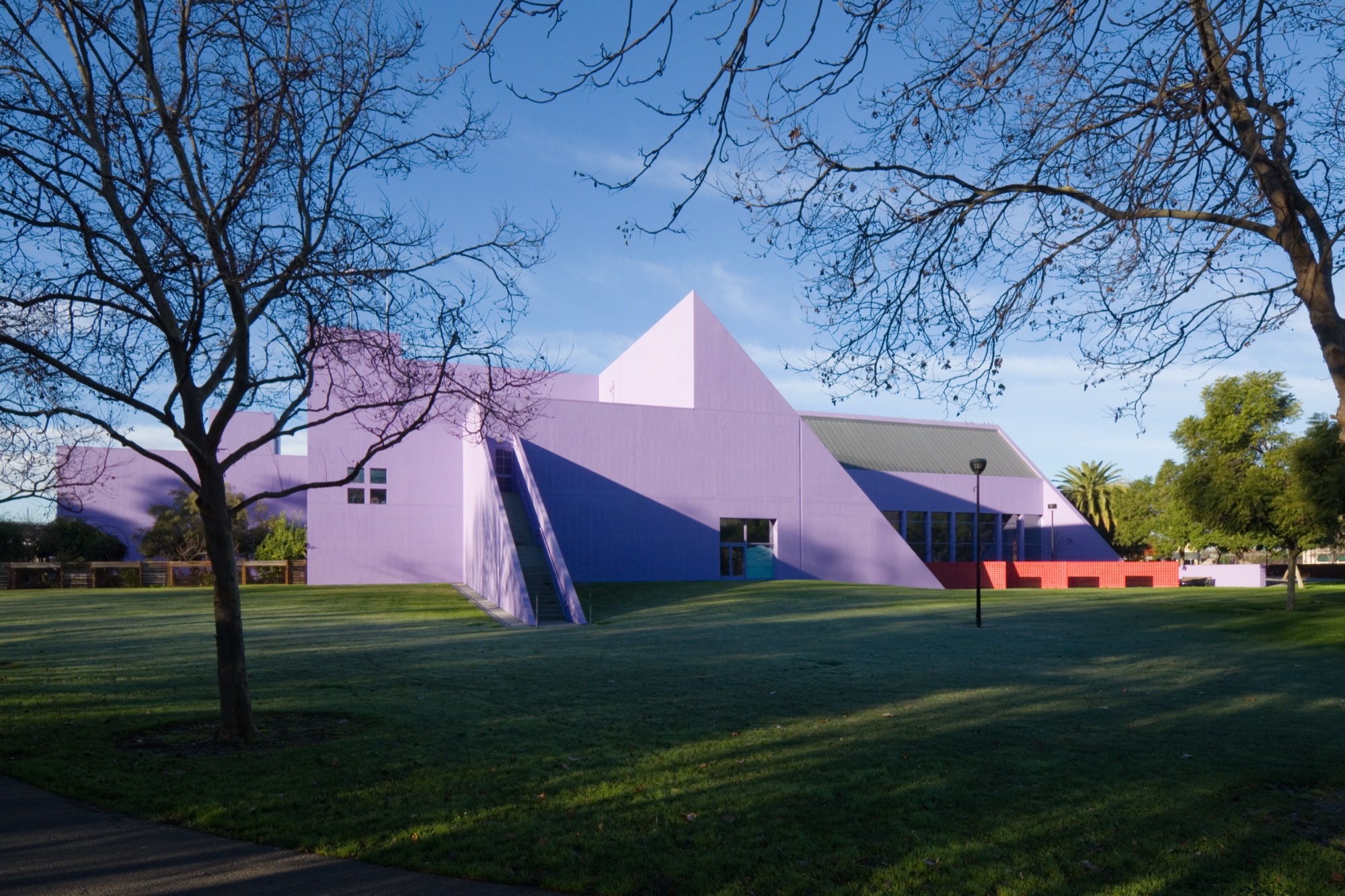 The lavender Children's Discovery Museum of San Jose, a low geometric building with a triangular roof, on a lawn under bare winter trees.