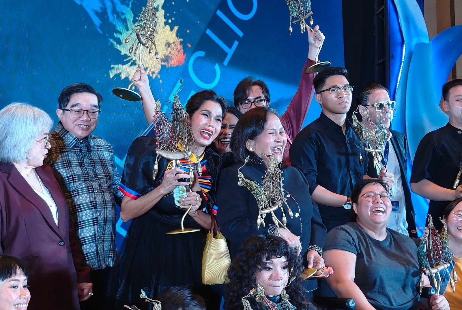 Pokwang and Ruby Ruiz hold matching Balanghai trophies on the Cinemalaya awards stage, laughing with other winners behind them.
