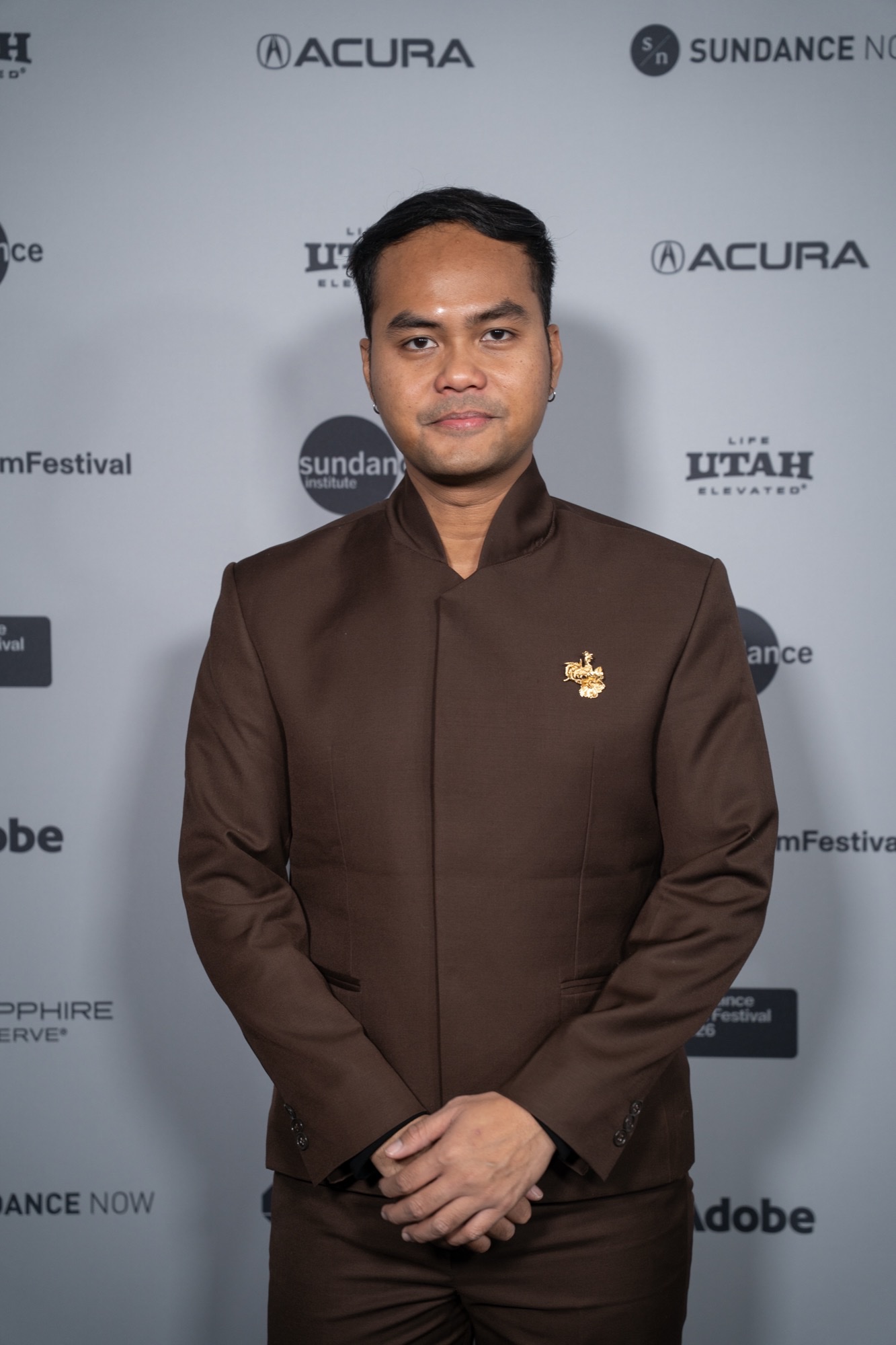 Wregas Bhanuteja at the 2026 Sundance premiere of Para Perasuk, also called Levitating, in a brown suit with his hands clasped, against a Sundance step-and-repeat.