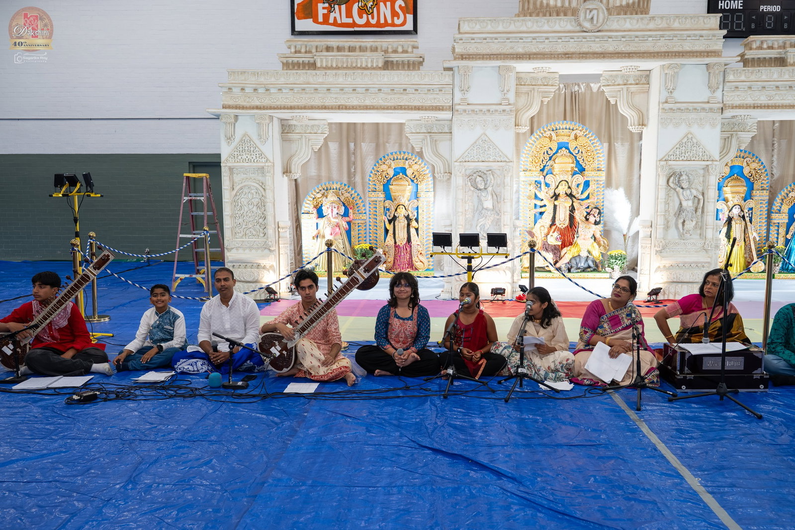 Musicians and singers seated on a blue floor in front of a carved white Durga Puja pandal with Goddess Durga and accompanying deities at a Dakshini Durgotsav.