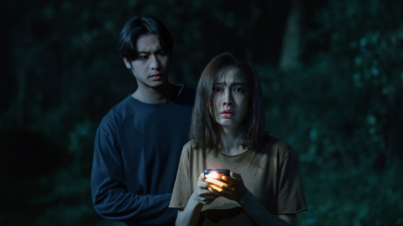 Natapohn Phornprapha as Wan holds a flashlight in dark woods, with Jespipat Tilapornputt as detective Pete standing behind her, in Netflix's Delusion.