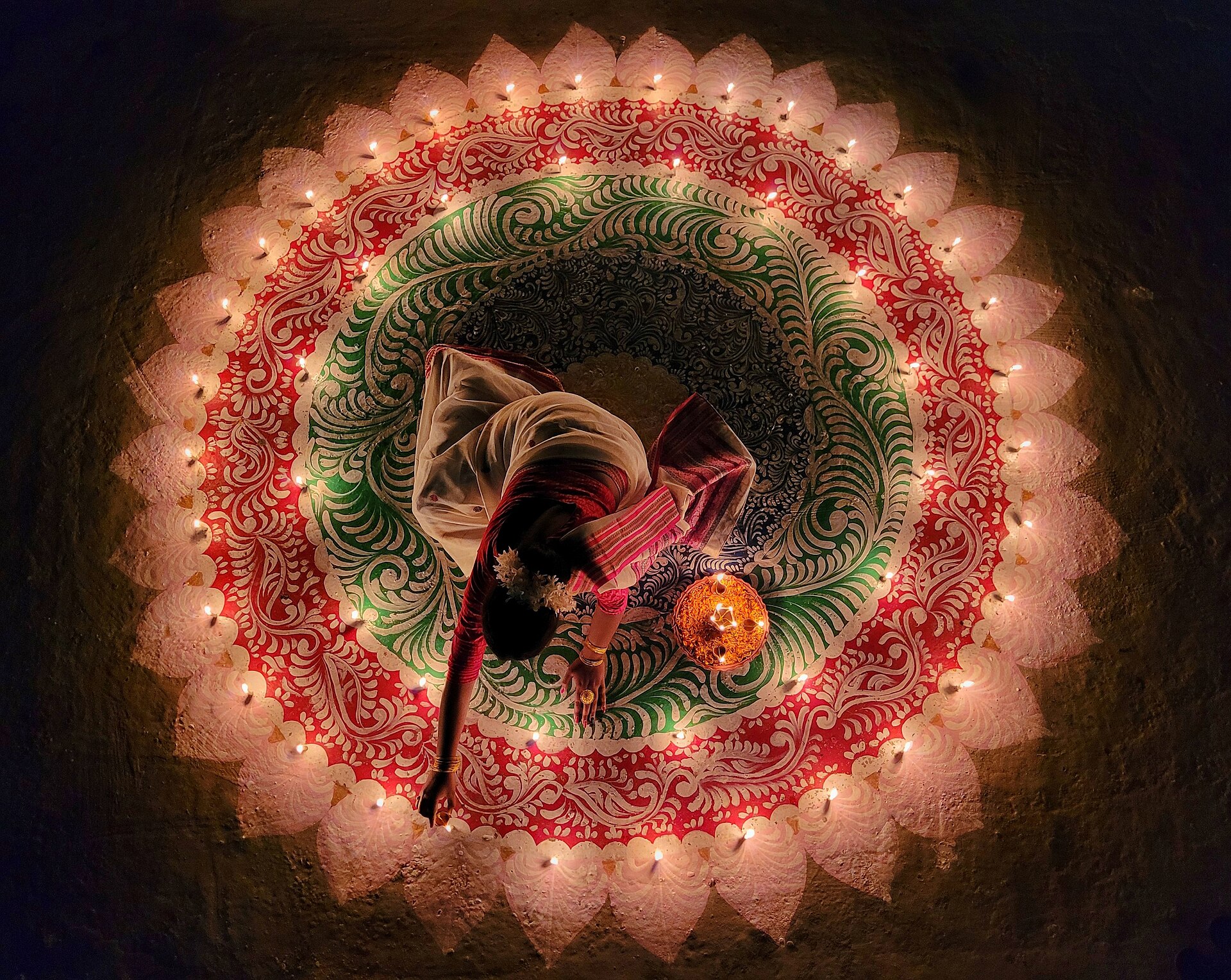 Context image: Woman in a white and red sari lighting diyas around an intricate rangoli pattern.