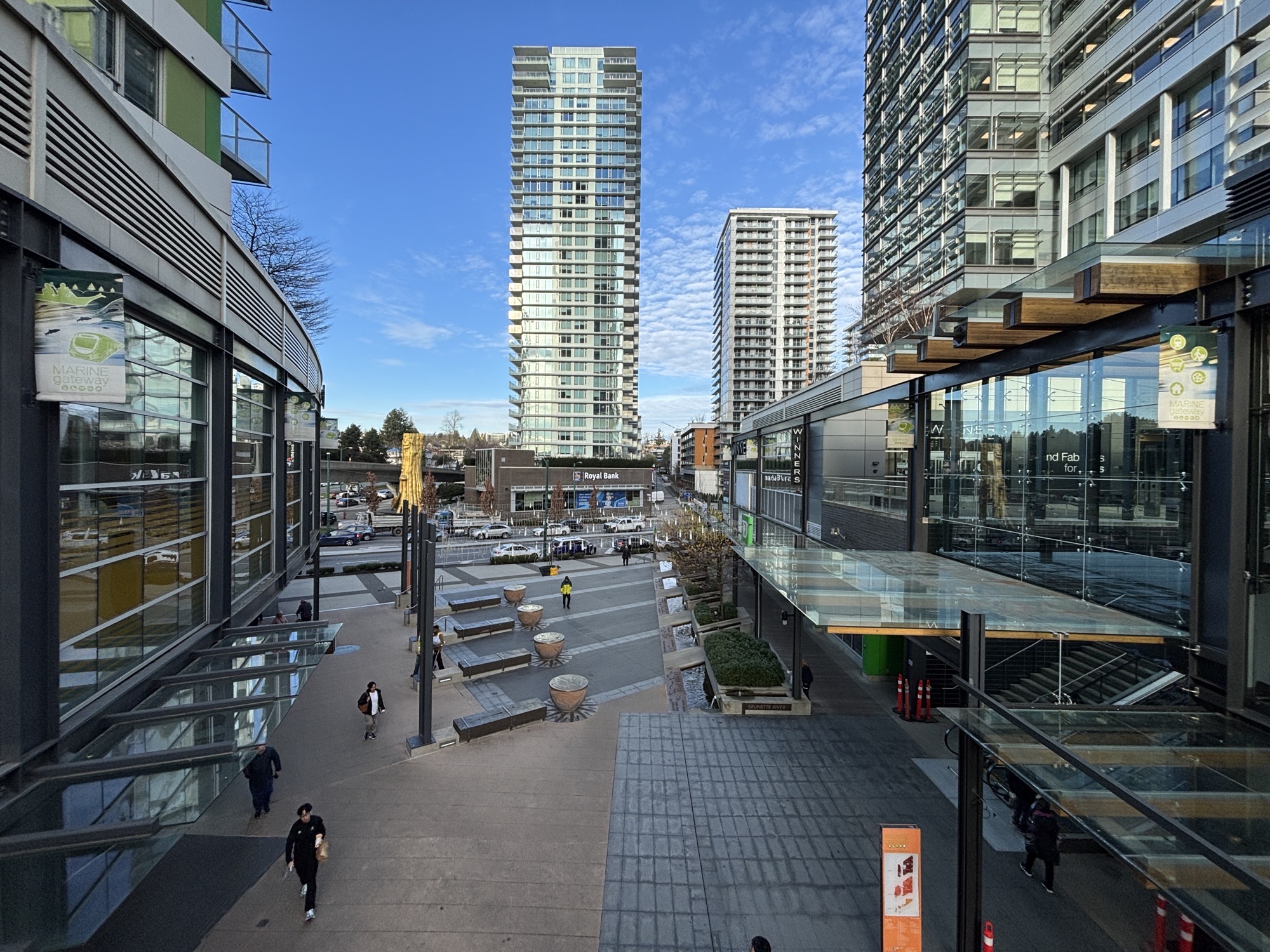 The Marine Gateway plaza in South Vancouver, with the mall sign, glass storefronts, and towers around an open pedestrian square.