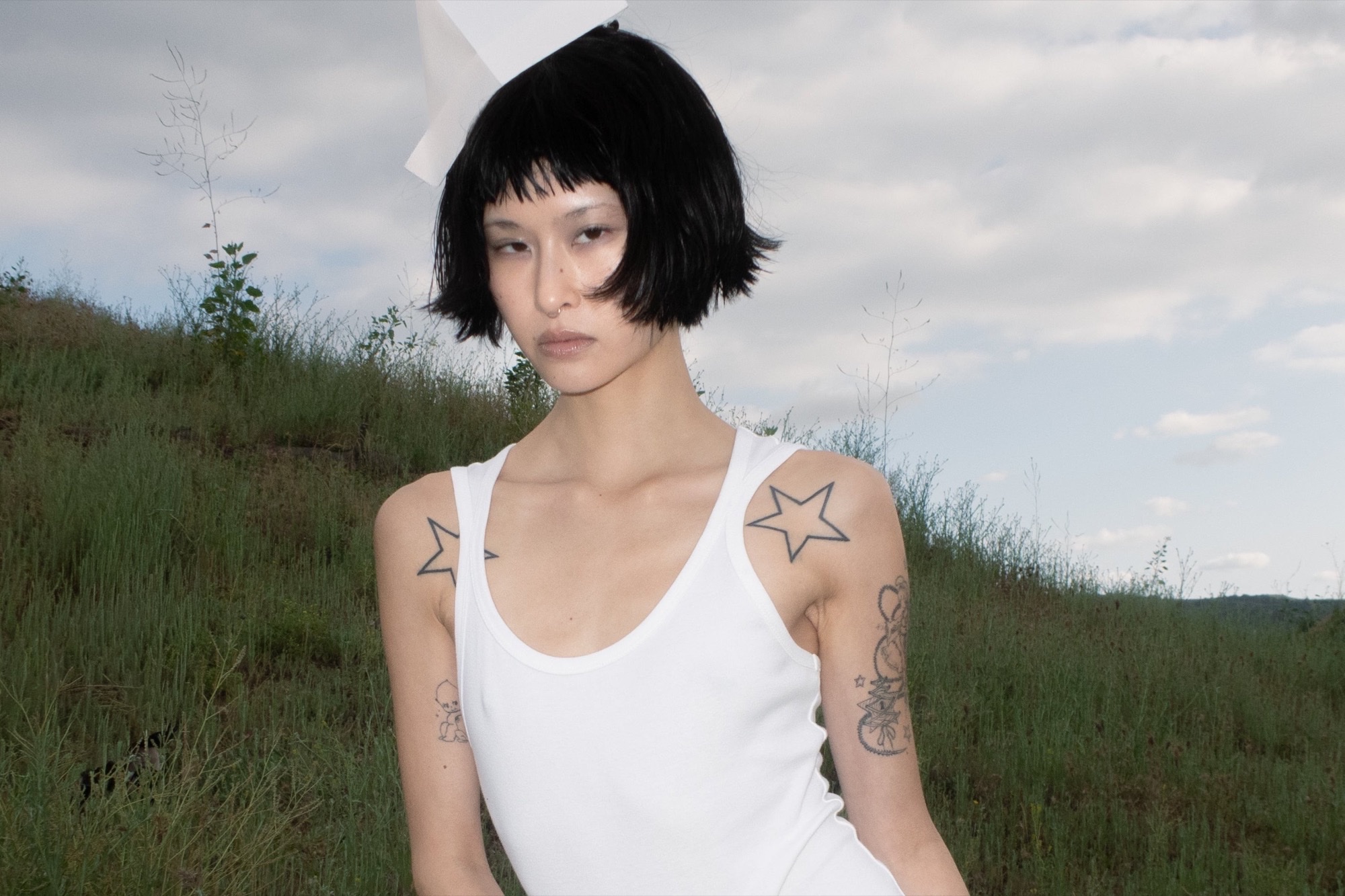 EMPATH founder Emily Arakawa in a white tank against a grassy hillside, short black bob and star tattoos visible on her shoulders.