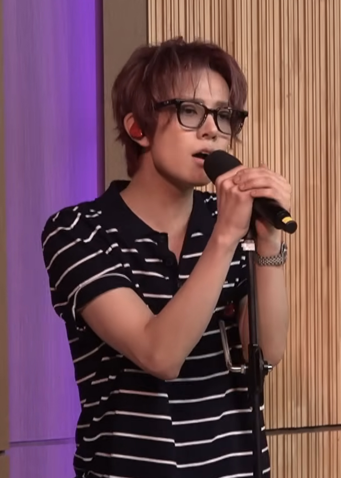 EVAN, formerly ENHYPEN's Heeseung, sings into a microphone on a live set while wearing glasses and a striped polo.