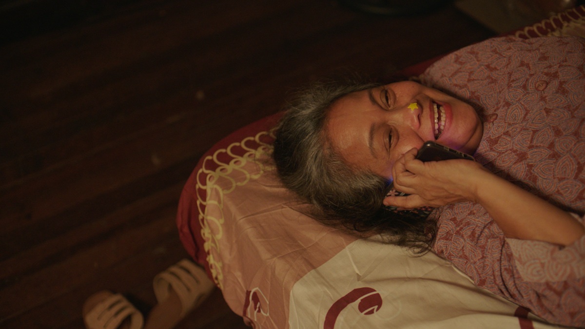 Ruby Ruiz as Biring in Status: Rejected, laughing on a phone in bed with a yellow star sticker on her nose.