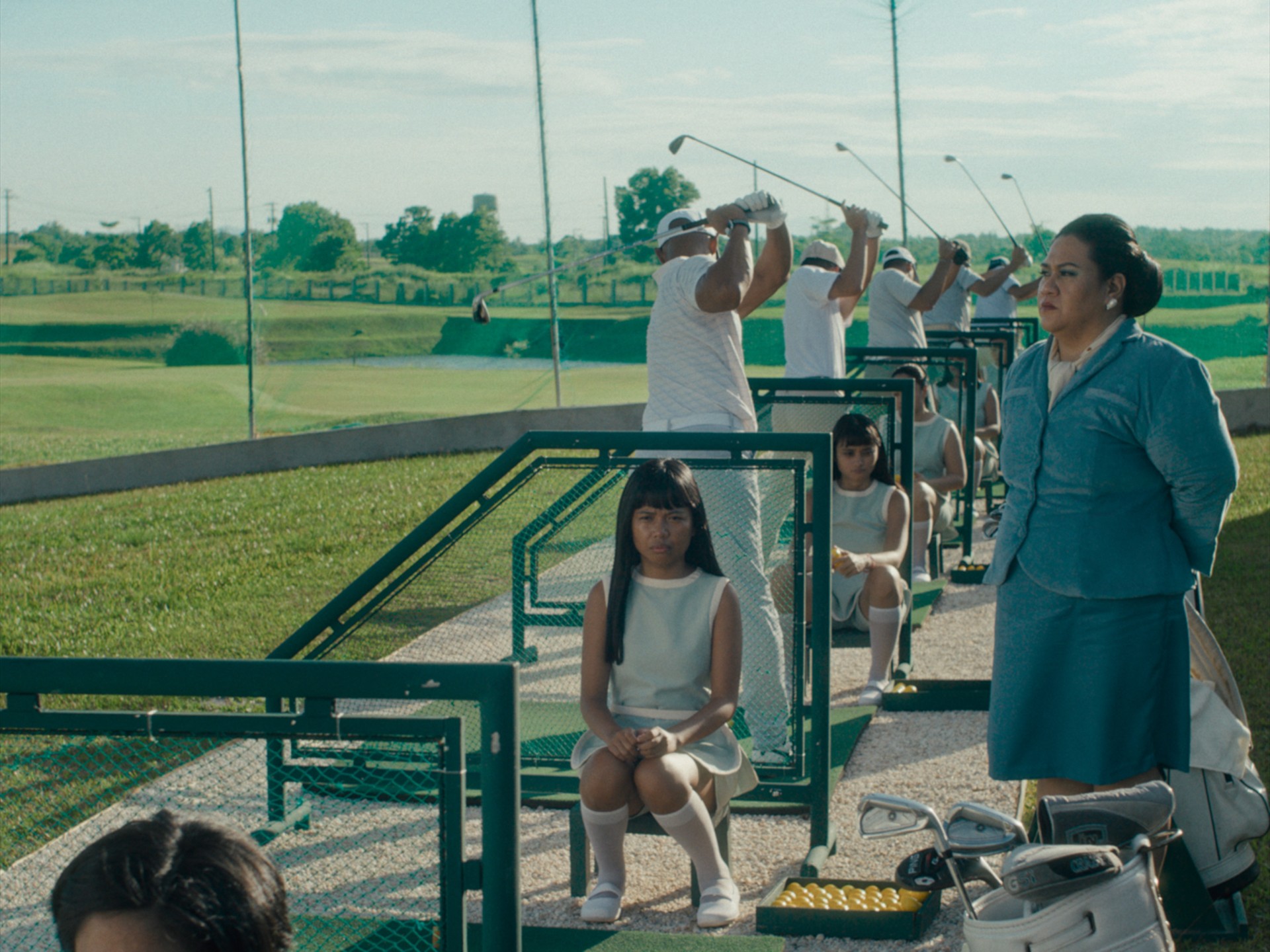Jorrybell Agoto as Isabel sits in a pale uniform at a driving range, while men in white swing golf clubs behind her.