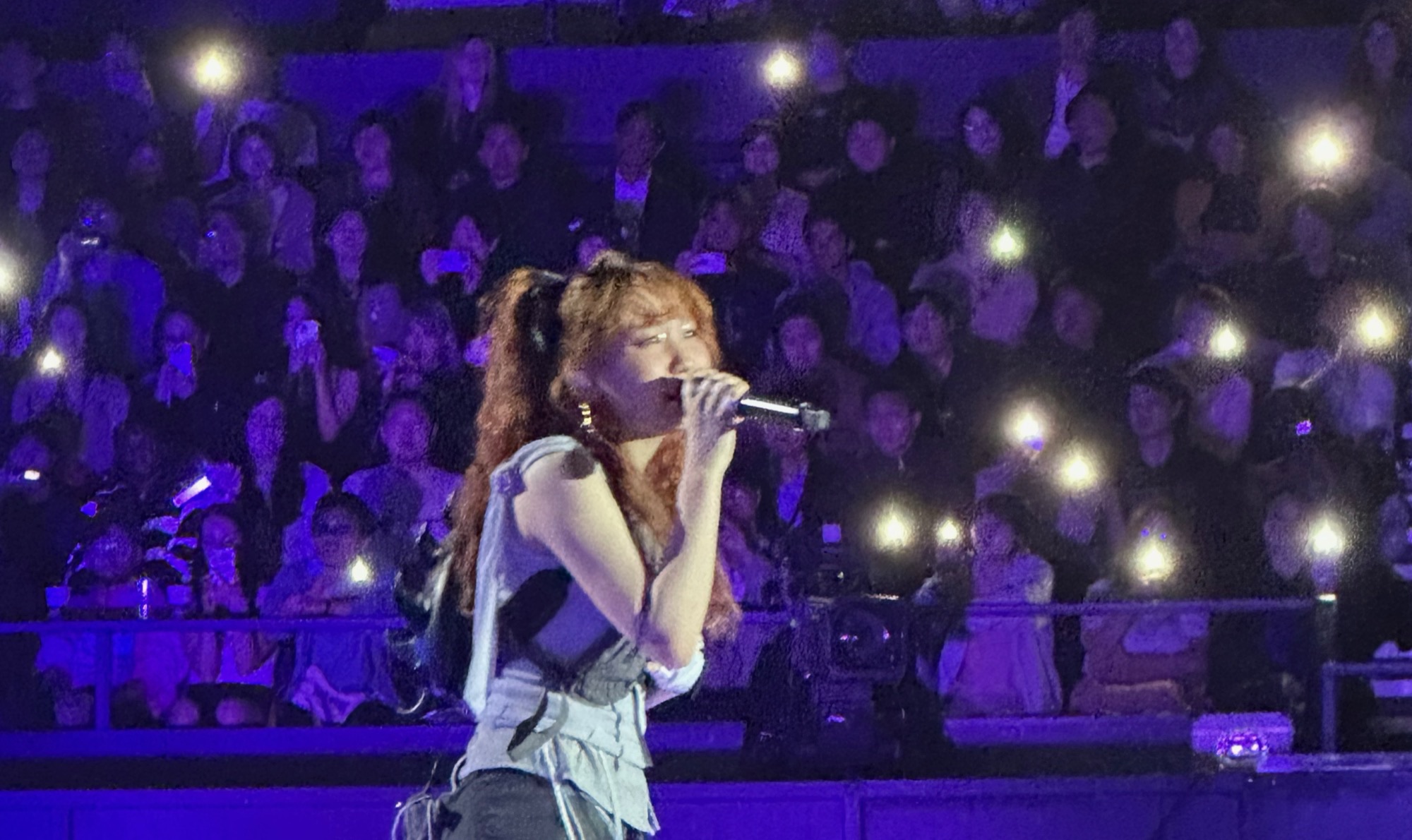 Lilas Ikuta of YOASOBI singing into a microphone on a purple-lit stage, with a phone-light crowd behind her.