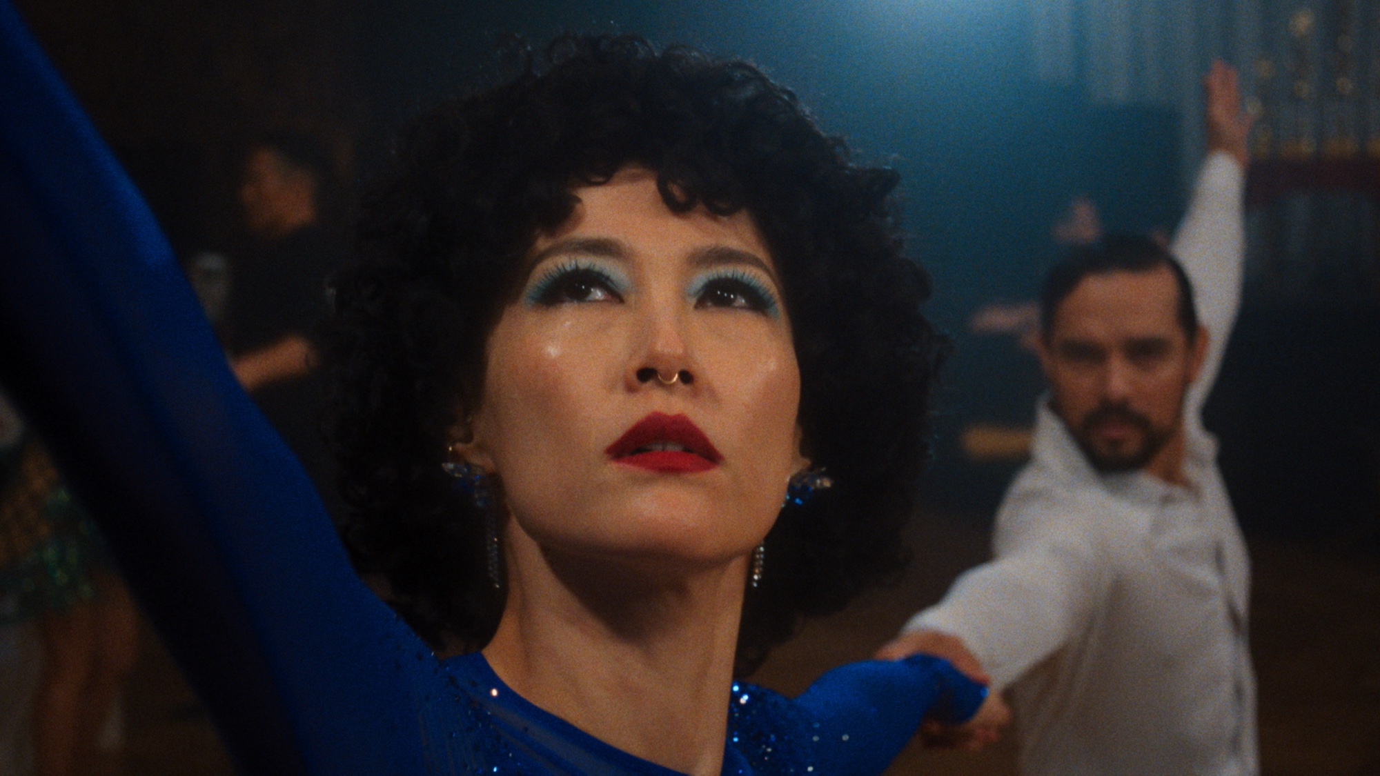 Rinko Kikuchi as Haru, in a blue sequined dress with a curled wig, dances in a ballroom, with a partner in a white shirt behind her.