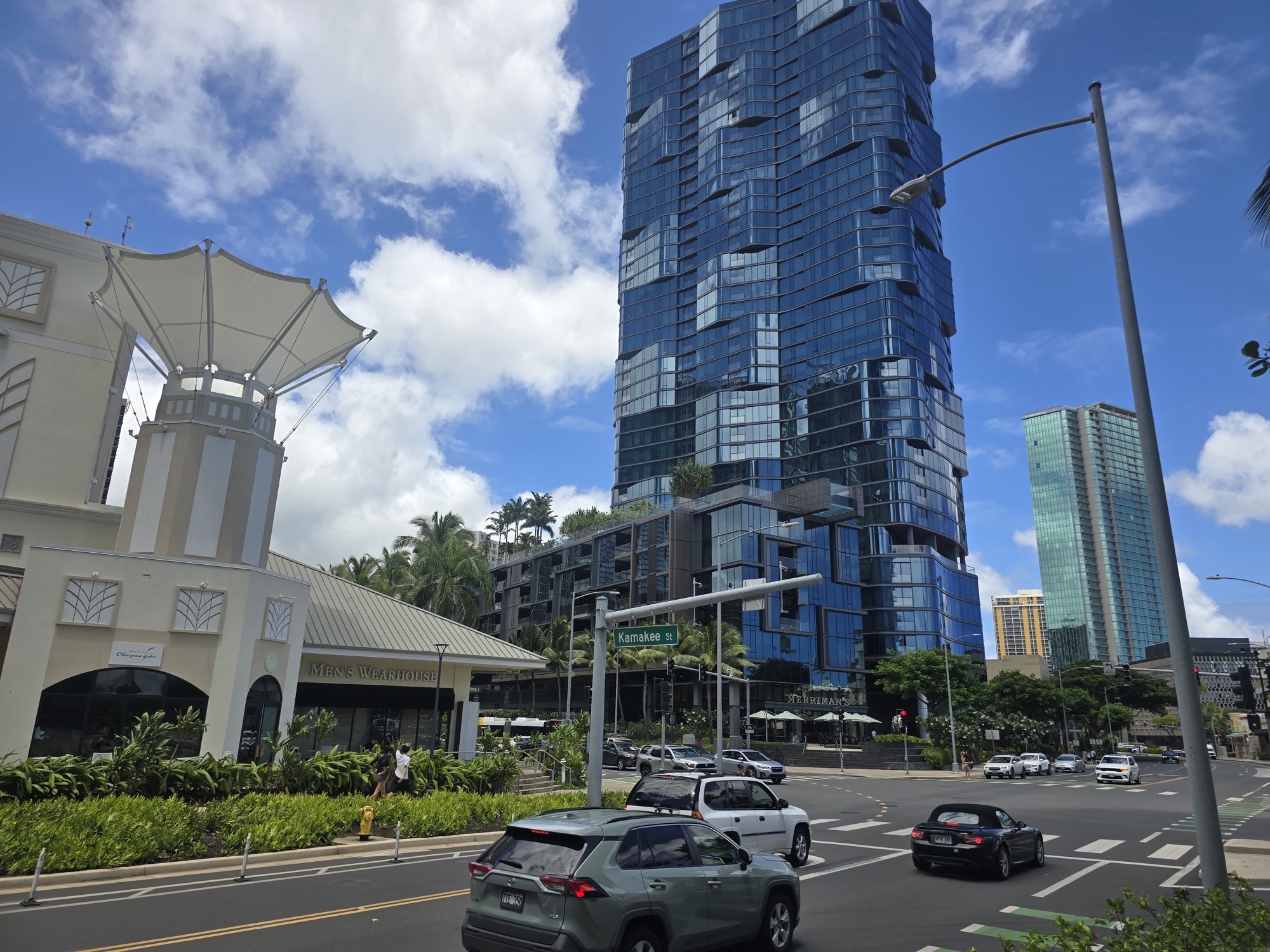 Kamakee Street corner in Honolulu's Kakaʻako, with glass condo towers, tropical landscaping, and daytime traffic under a bright sky.