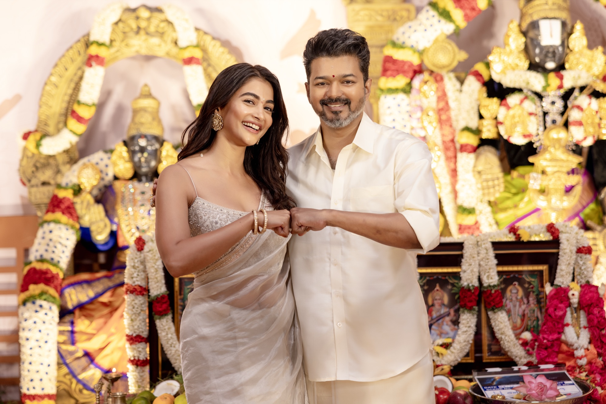 Vijay and Pooja Hegde fist-bumping at the Jana Nayagan puja with flower-garlanded temple statues and a film clapperboard in frame.