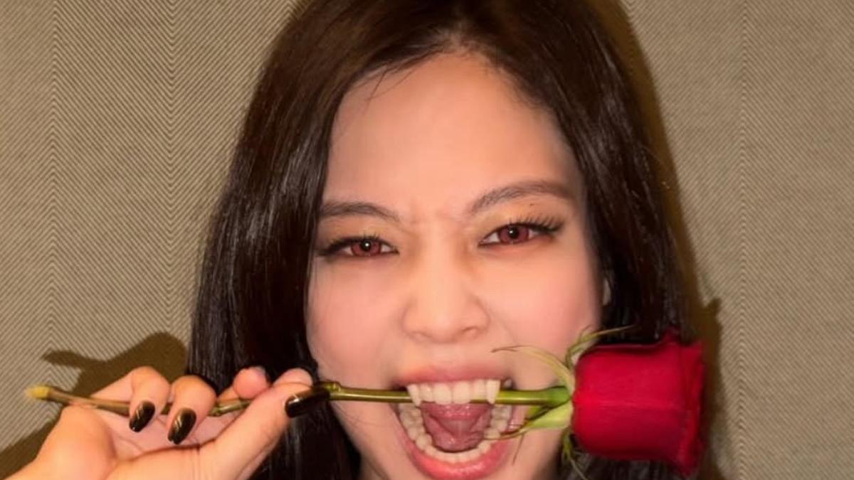 Jennie with red contact lenses and fangs bites the stem of a red rose, one hand holding the stem near her mouth.