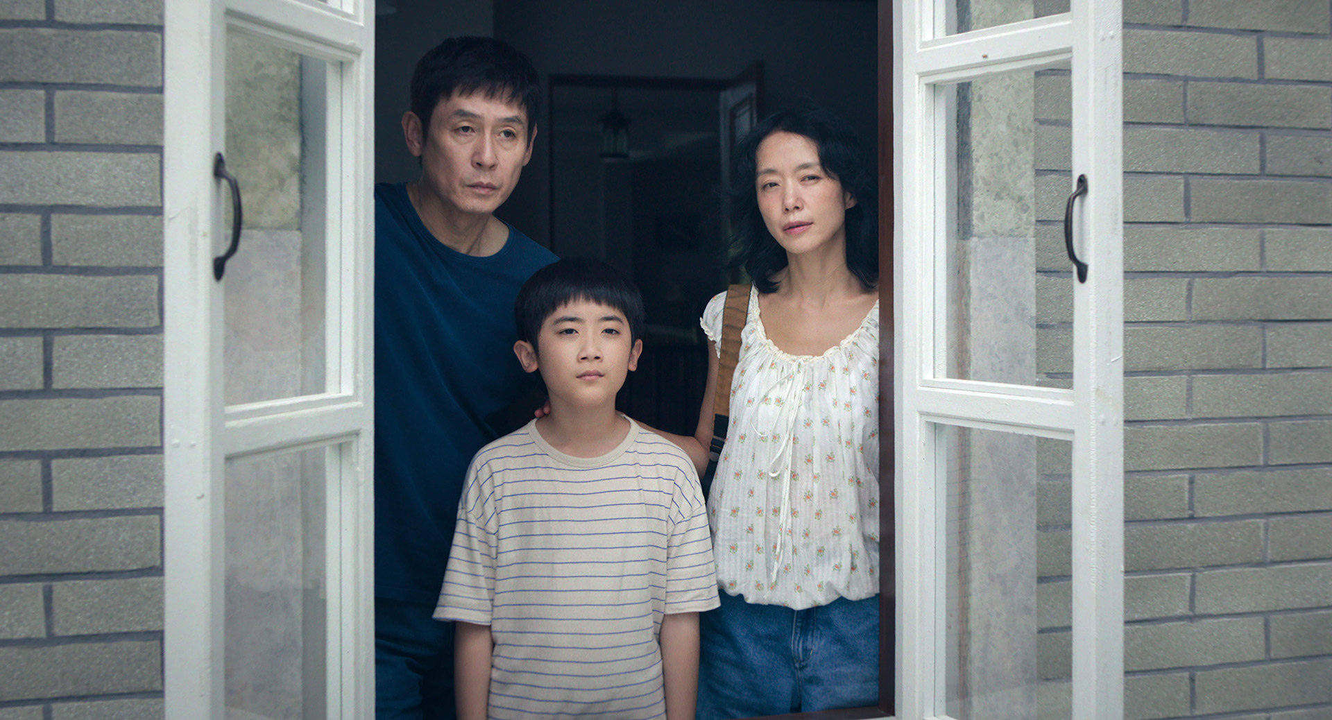 Jeon Do-yeon as Mi-ok stands in a doorway in a white blouse, beside her son in a striped shirt and Sul Kyung-gu in a blue t-shirt.