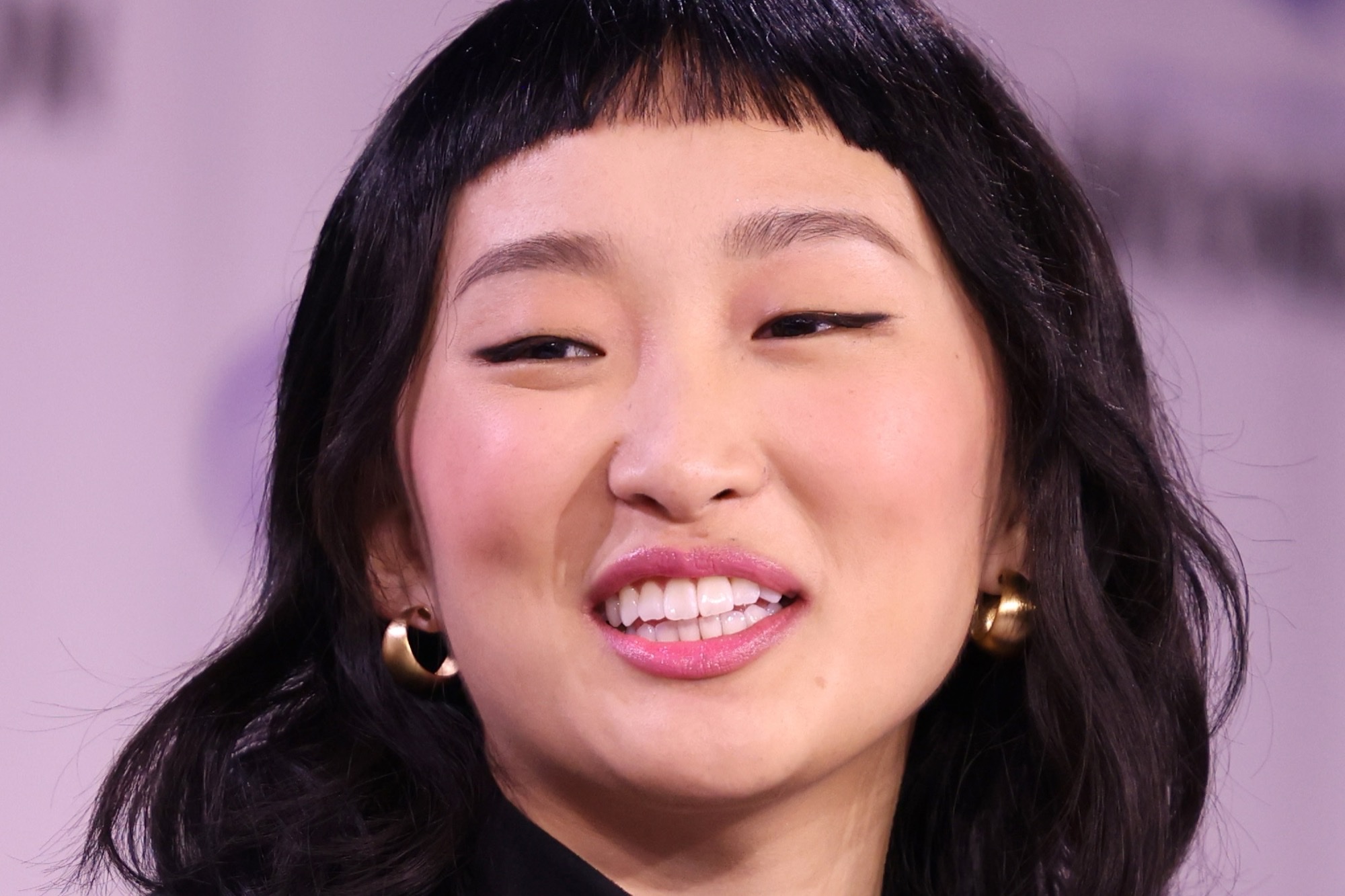 Ji-young Yoo smiling at a press event, dark wavy hair with micro bangs and gold hoop earrings.