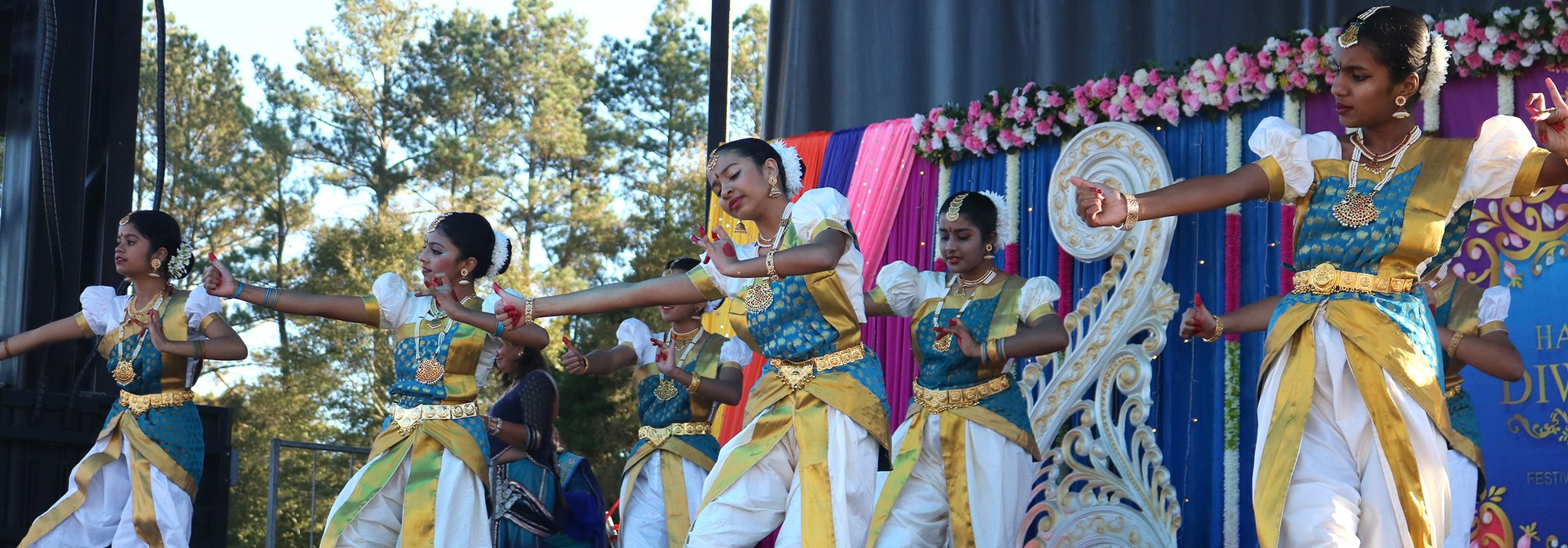 Young dancers in classical costume perform on an outdoor Diwali stage with a Happy Diwali banner behind them.