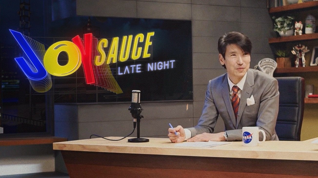 JoySauce founder Jonathan Ng Sposato hosts JoySauce Late Night at a studio desk with the show logo on a screen behind him.