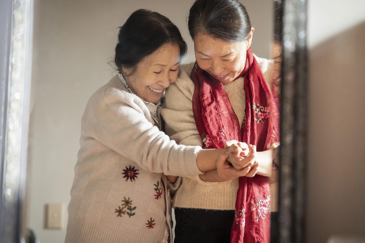 Kim Hye-ja and Cha Mi-kyung in Still Shining, smiling and holding hands in a sunlit doorway, one in a cream cardigan and one in a red scarf.