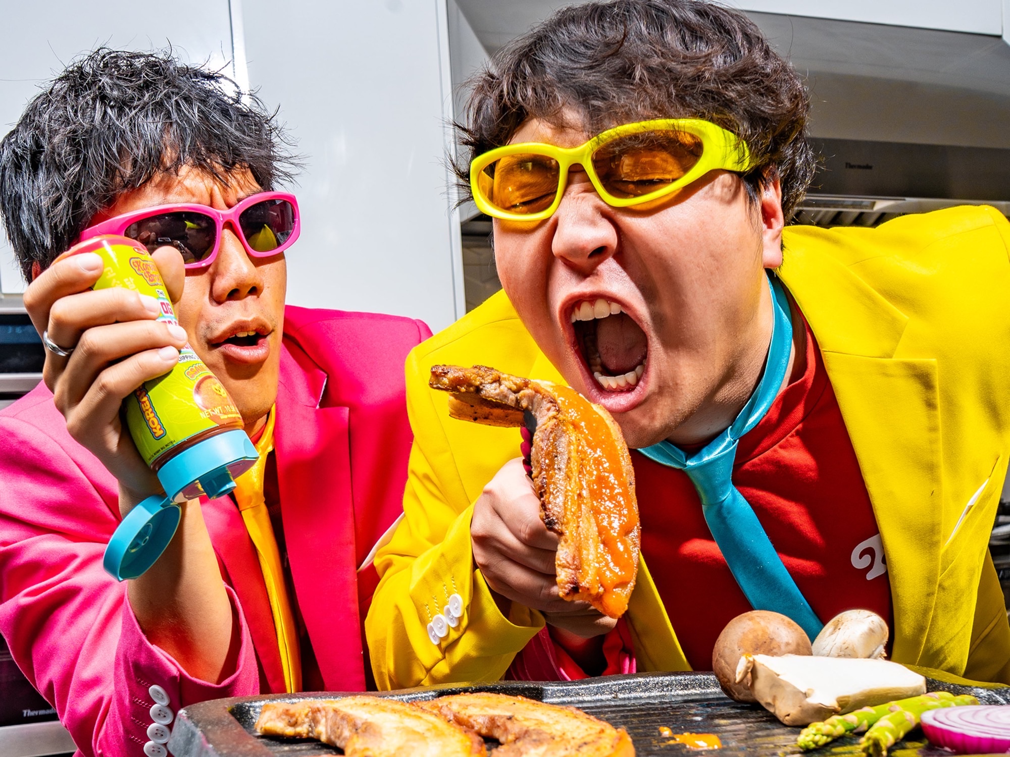 Korean Bros co-founders in bright suits sharing grilled pork belly and sauce in a kitchen.
