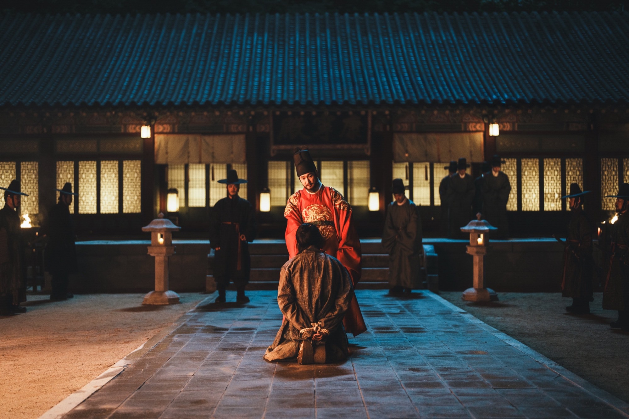 Cho Seung-woo as the king in a red royal robe stands over a kneeling prisoner in a night palace courtyard in Netflix's The East Palace.
