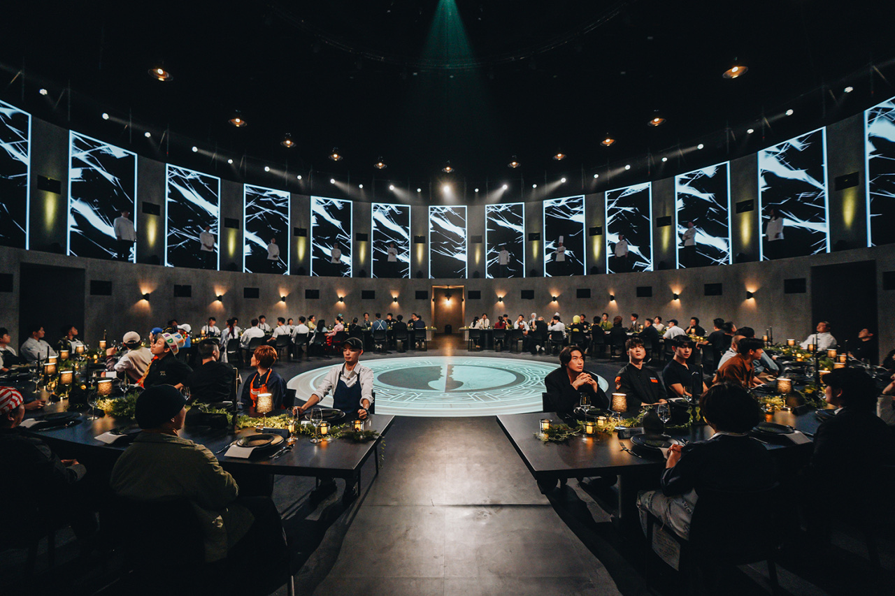 Contestants seated around a circular studio set for Netflix’s Culinary Class Wars, with theatrical lighting and a glowing center emblem on the floor.