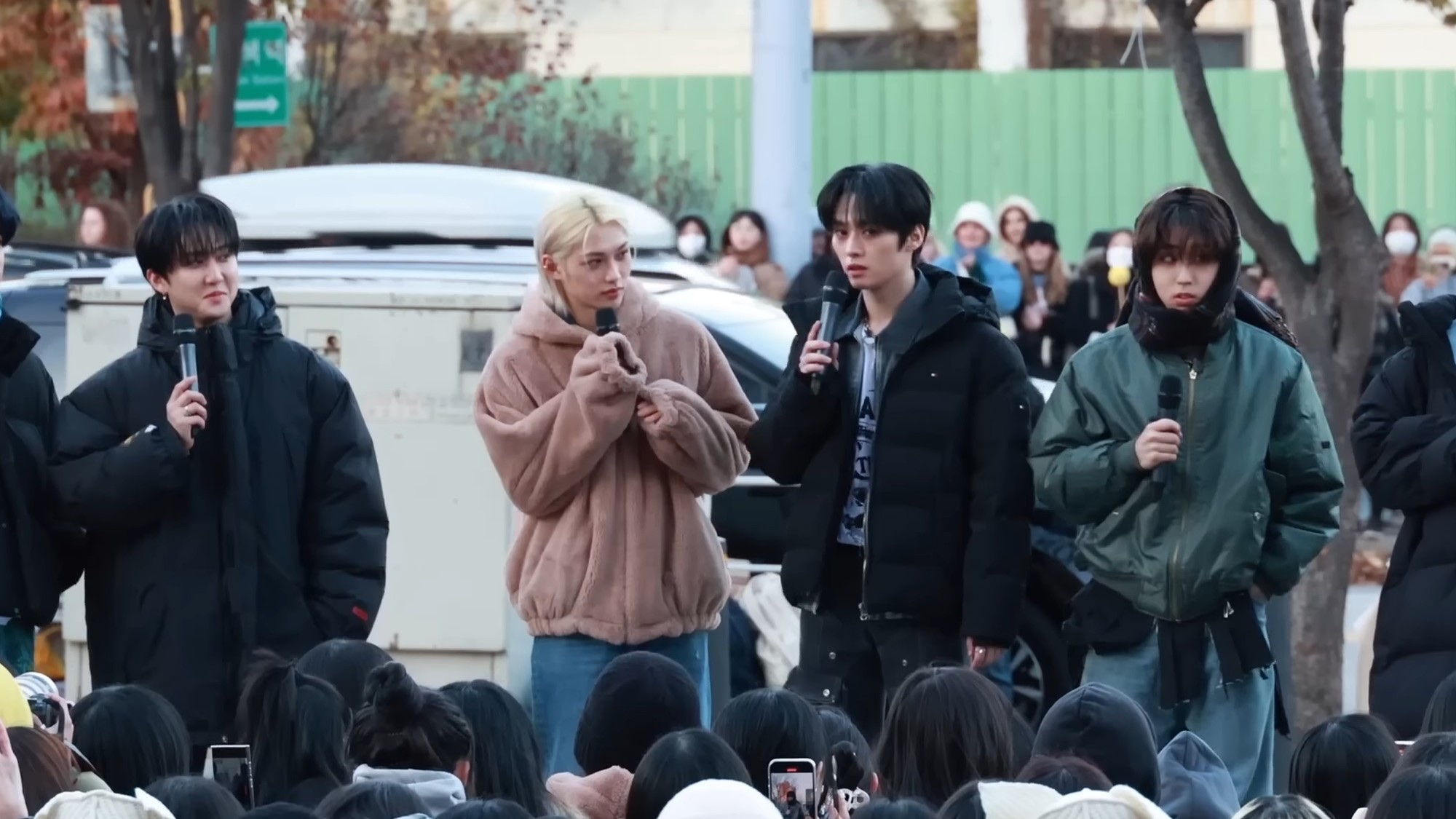 Four Stray Kids members in winter coats holding mics at an outdoor fan meeting, with phones raised in the crowd.