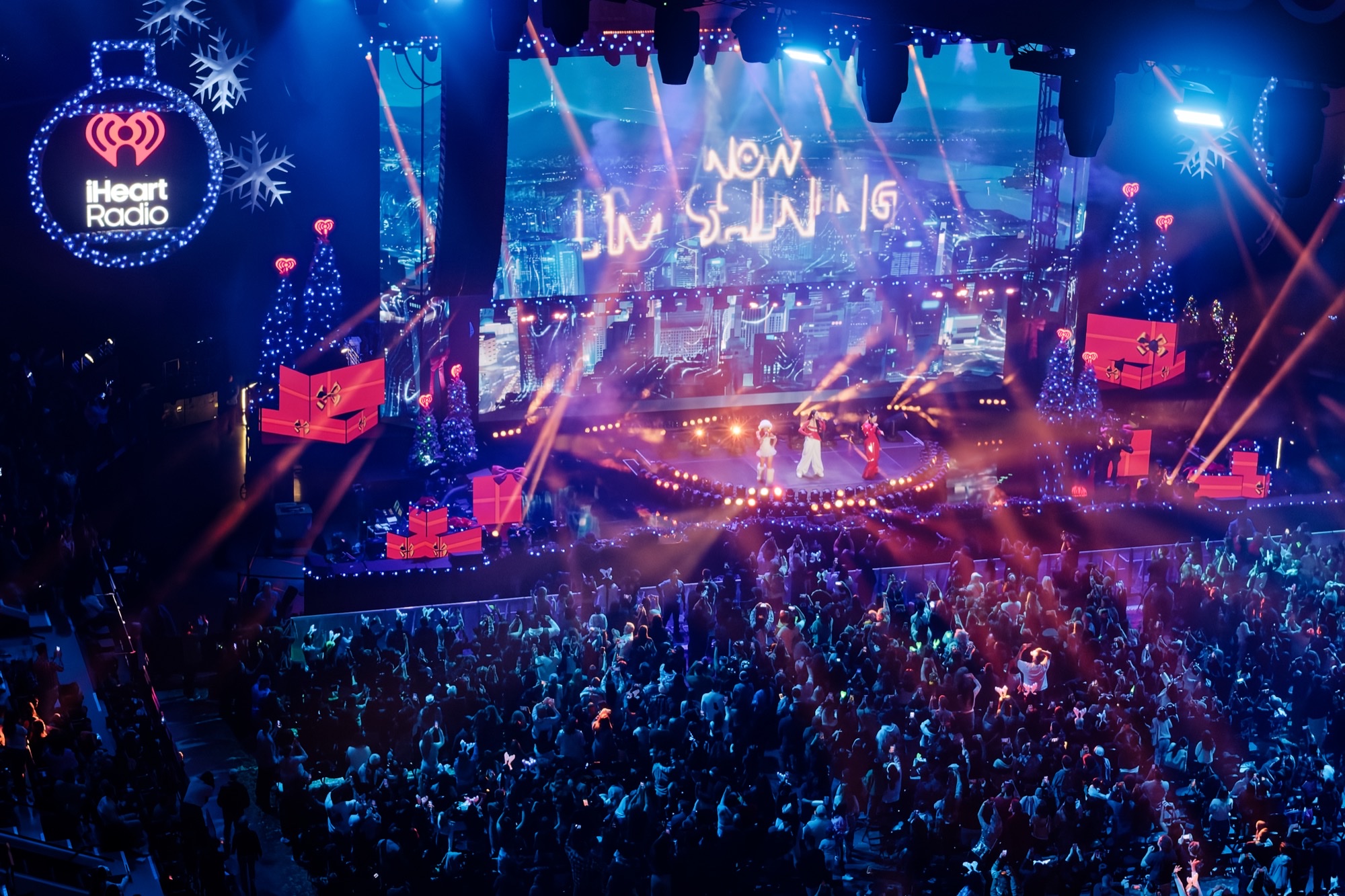 EJAE, Audrey Nuna, and REI AMI perform as HUNTR/X on the Jingle Ball stage under a NOW I’M SHINING screen, with a packed arena of lightsticks and phones.