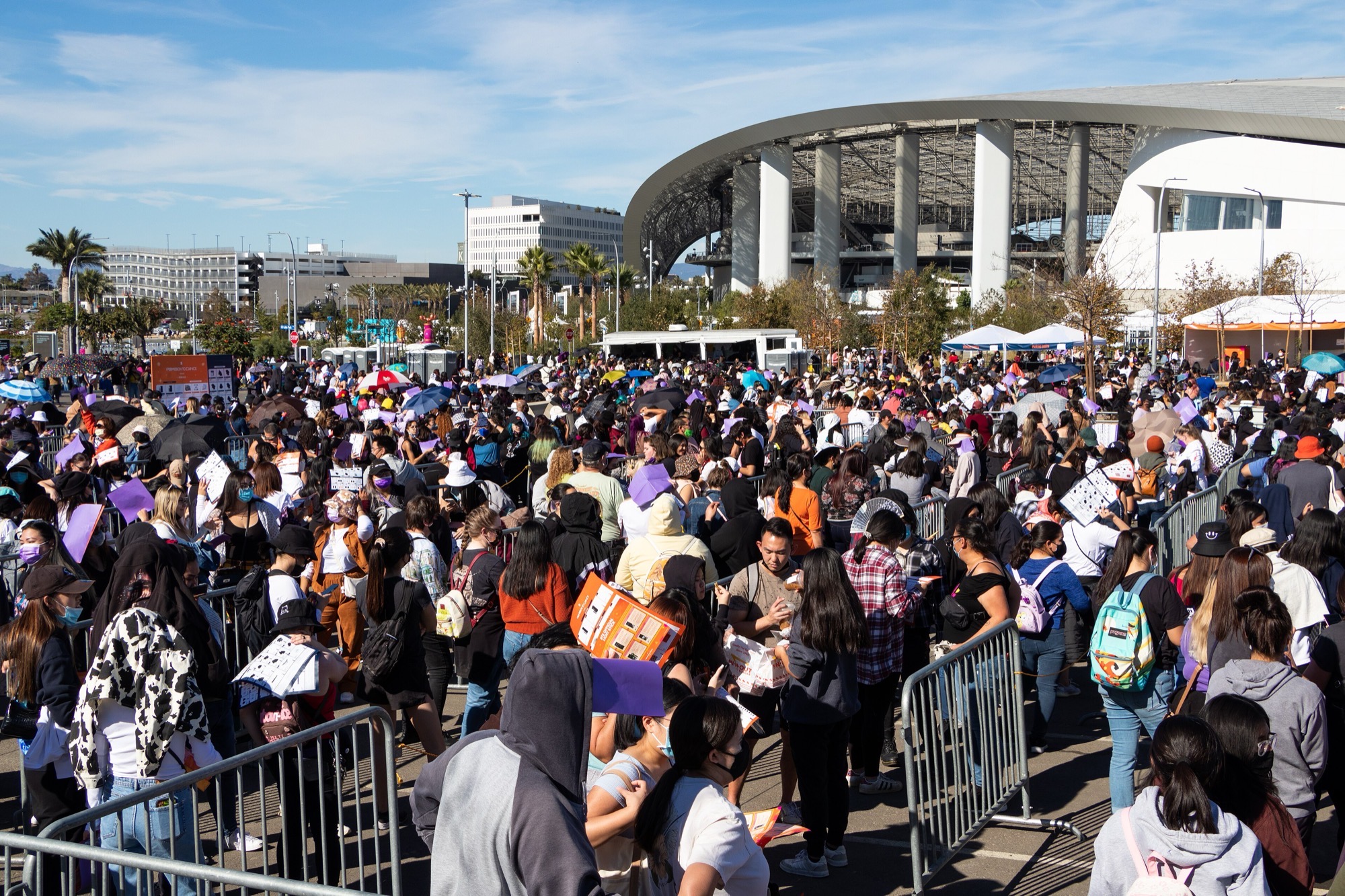 Crowds of BTS fans queued behind barricades outside SoFi Stadium in Inglewood, with purple umbrellas and white event tents under a clear sky.