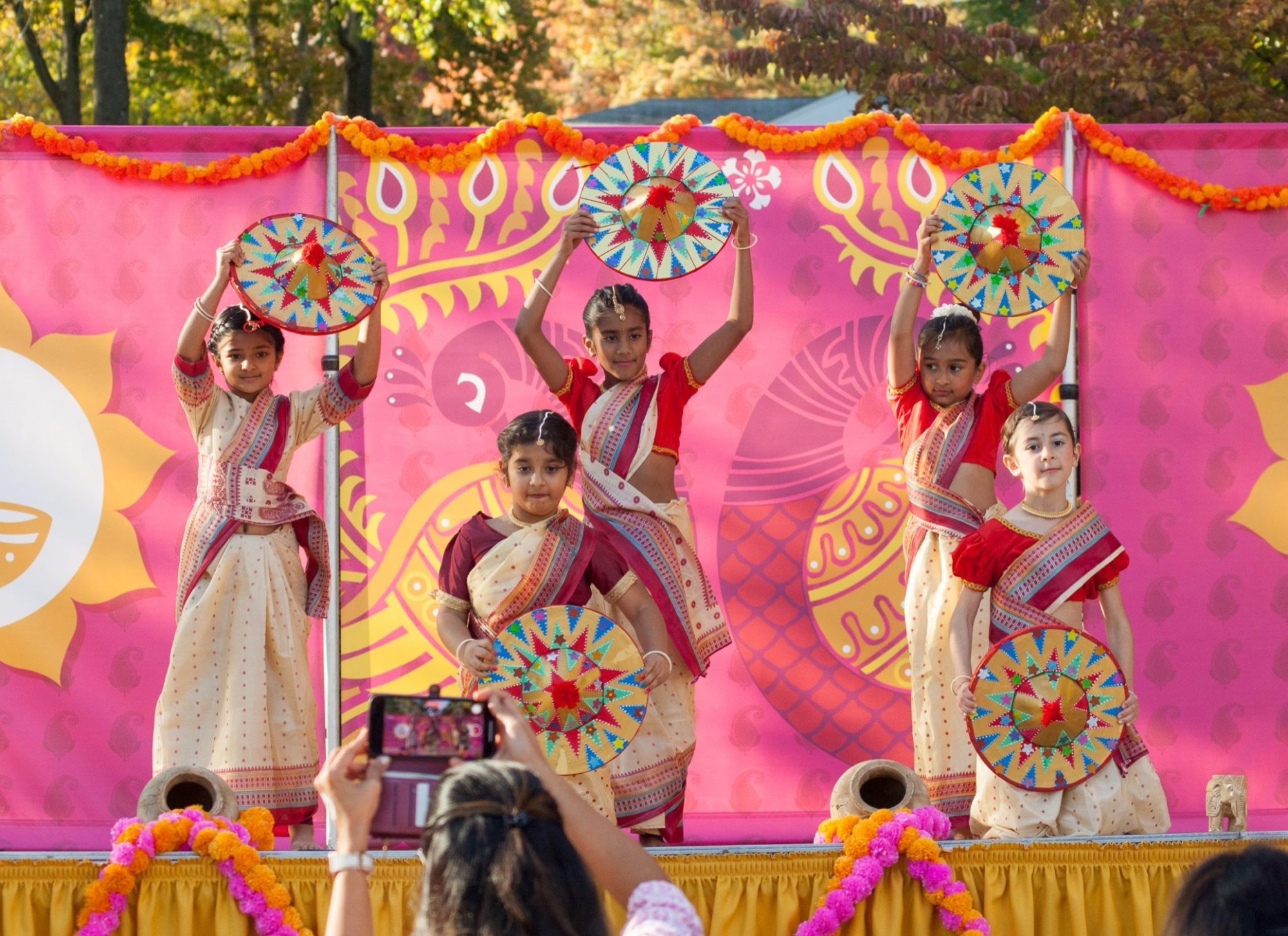 Young dancers in traditional Assamese dress hold colorful jaapi hats on an outdoor stage at a prior Light Up Diwali celebration, with marigold garlands and a pink backdrop behind them.