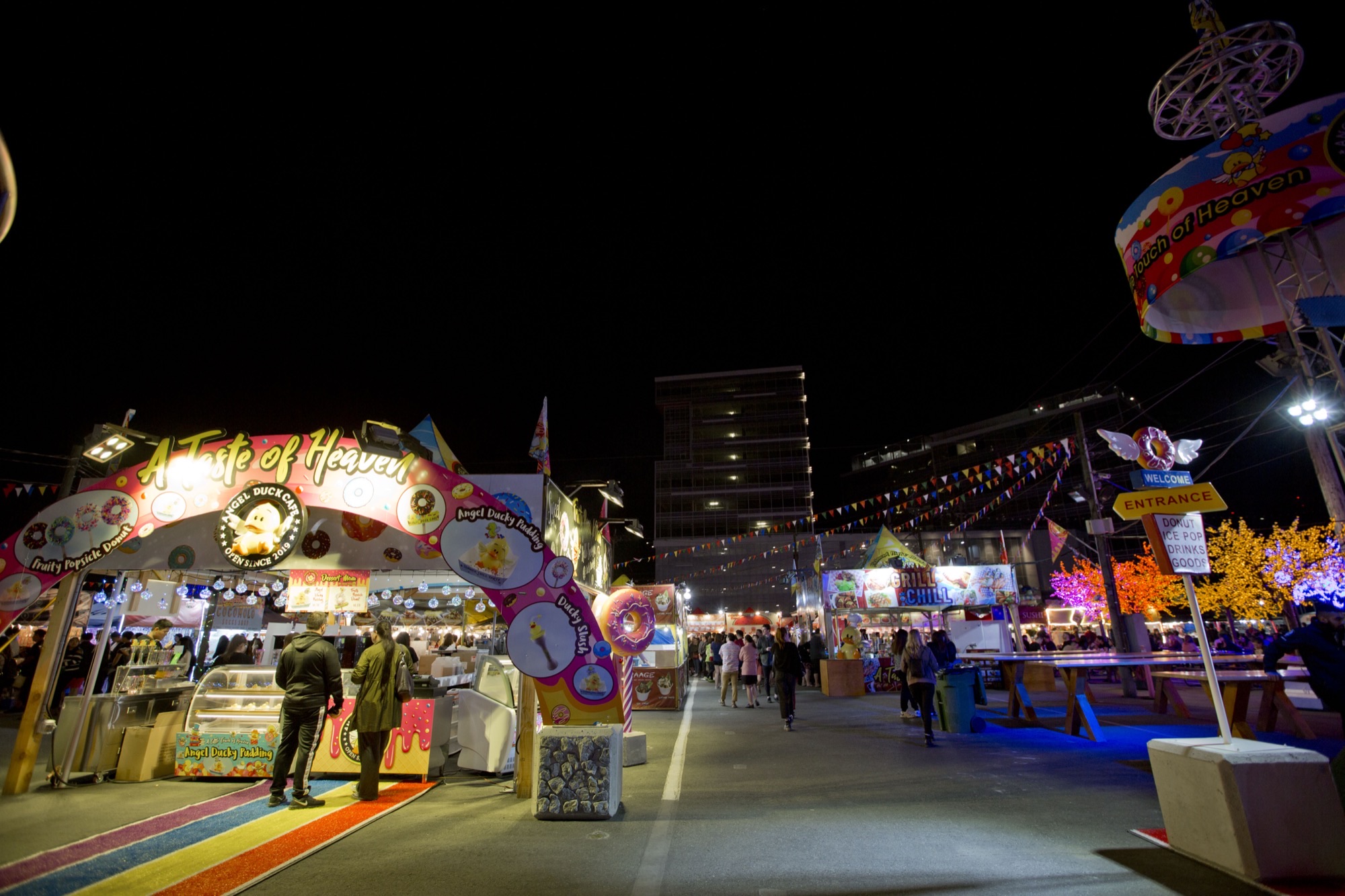 Lit food stalls and overhead flags at Richmond Night Market at night, with Angel Duck Cafe signage and a modern office building in the background.
