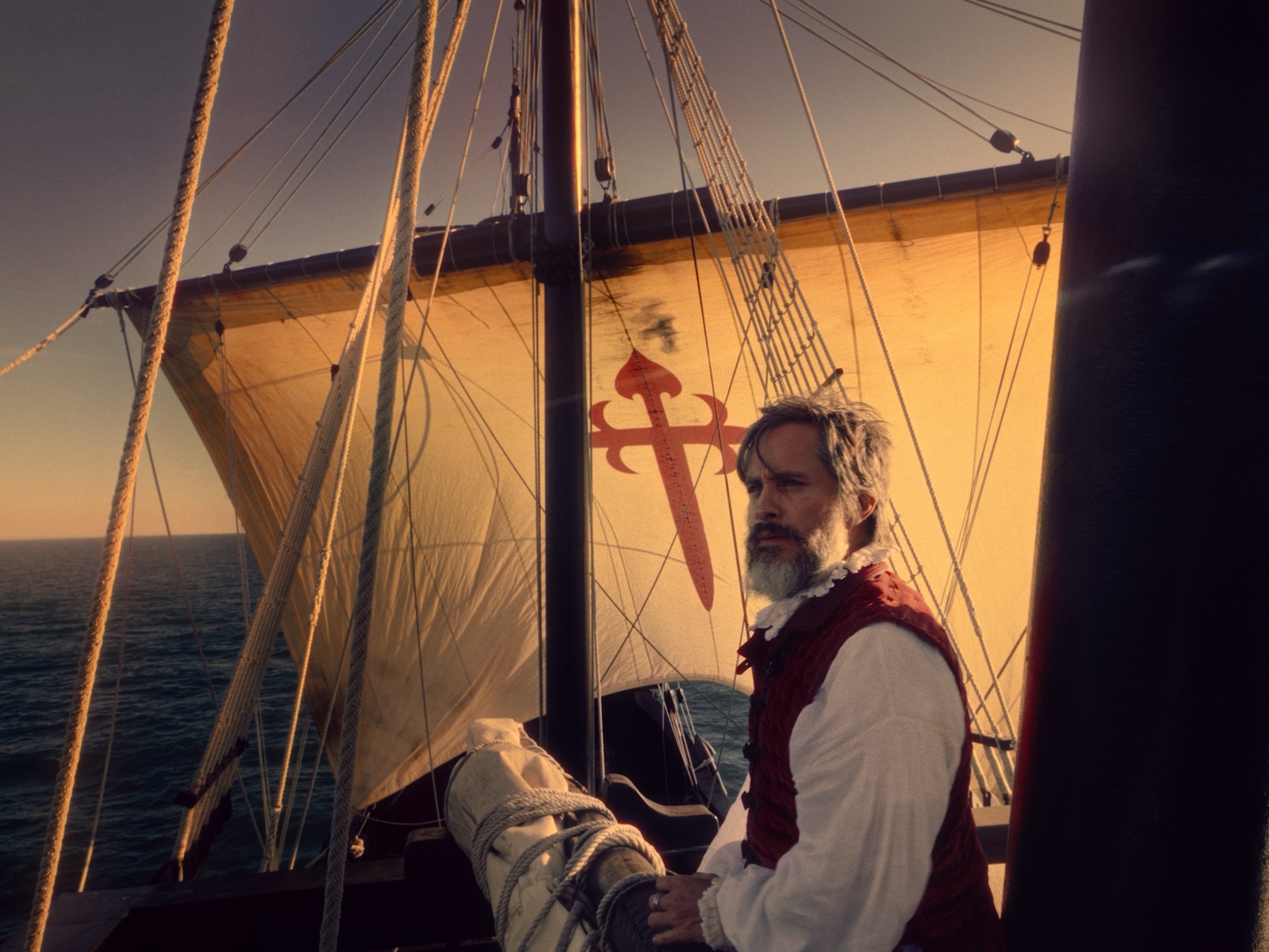 Gael García Bernal as Ferdinand Magellan stands at a ship's rail, with a red cross on the sail behind him, in Lav Diaz's Magellan.