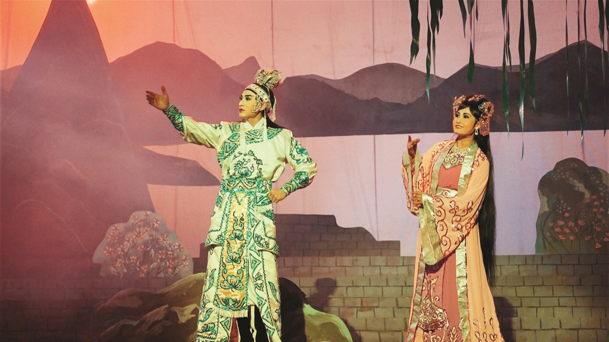 Two cải lương performers in ornate stage costume under painted sunset mountains, from Leon Le's Song Lang.