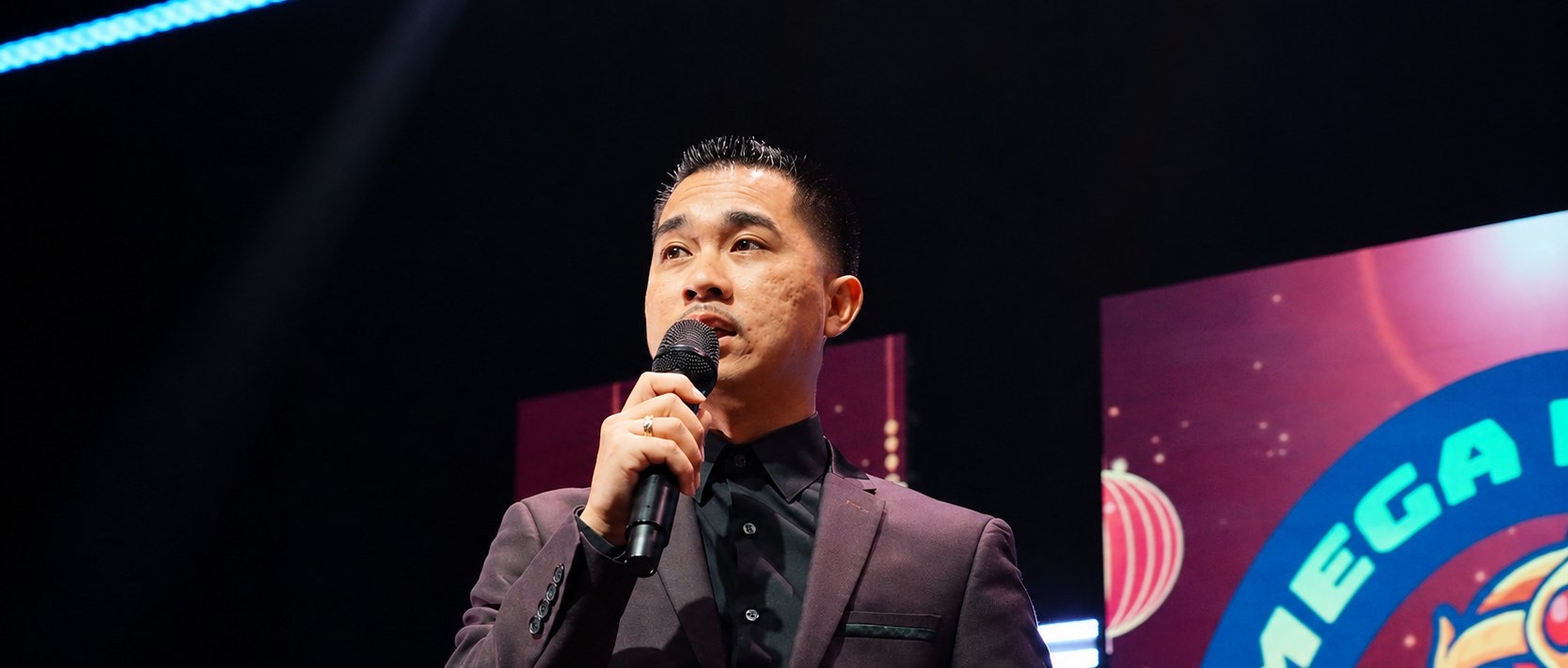 A man in a plum suit speaks into a microphone on a Mega Night Market stage, with the festival logo on a screen behind him.