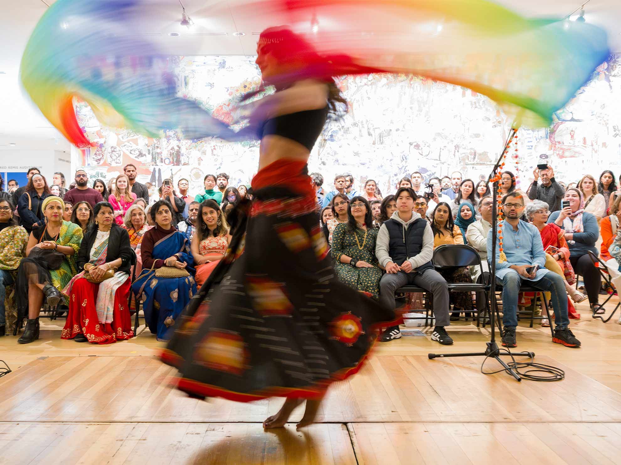 A dancer spins a rainbow veil on a wooden stage inside the Museum of Fine Arts, Boston, with seated visitors watching from gallery chairs.