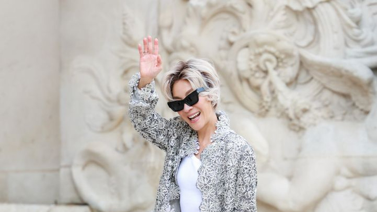 Michelle Yeoh waving outside Chanel's Paris haute couture show in a platinum blonde bob and embroidered Chanel skirt suit.