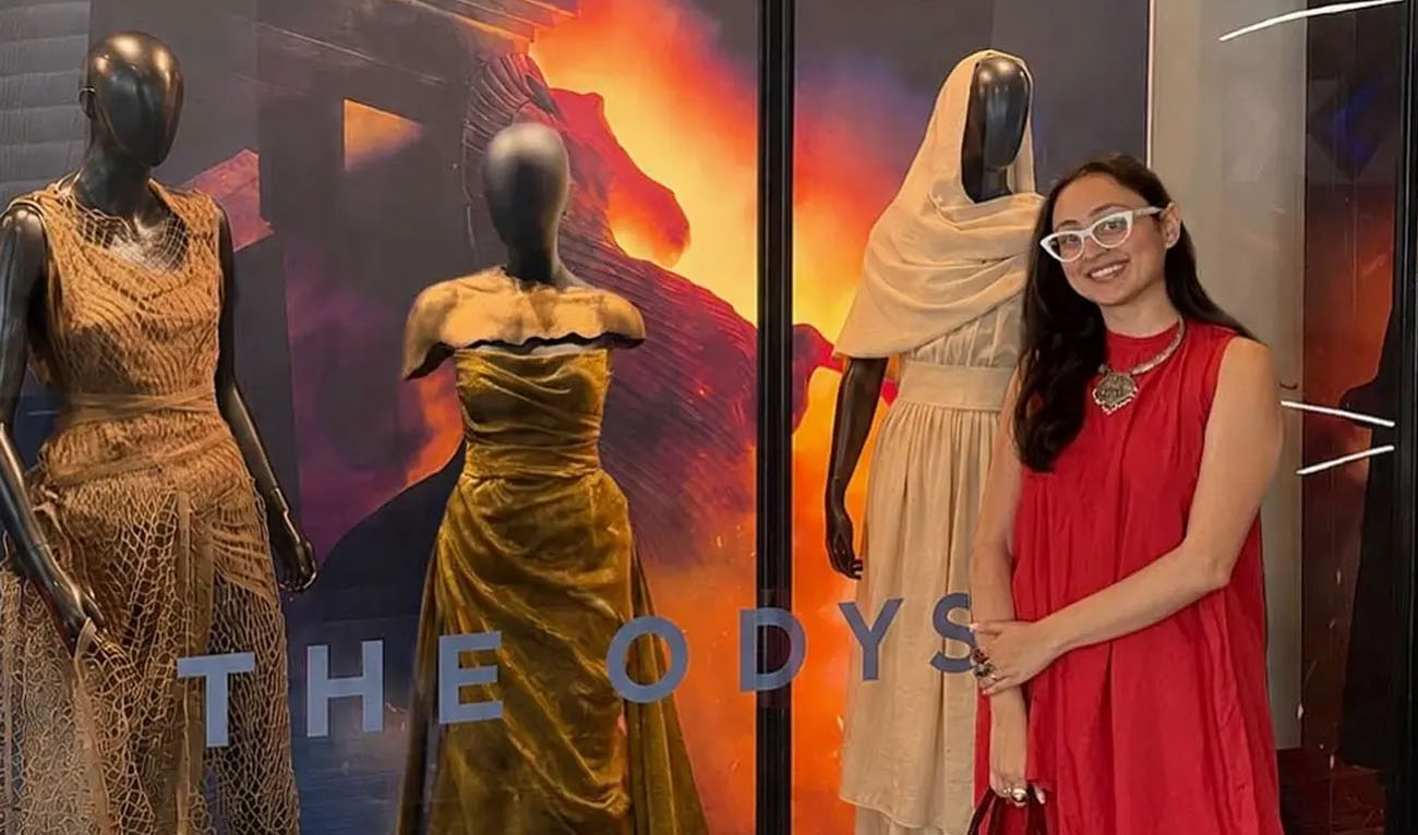 Misha Japanwala standing beside Odyssey costume mannequins, including a jagged bronze torso piece over a gold draped gown under THE ODYSSEY branding.