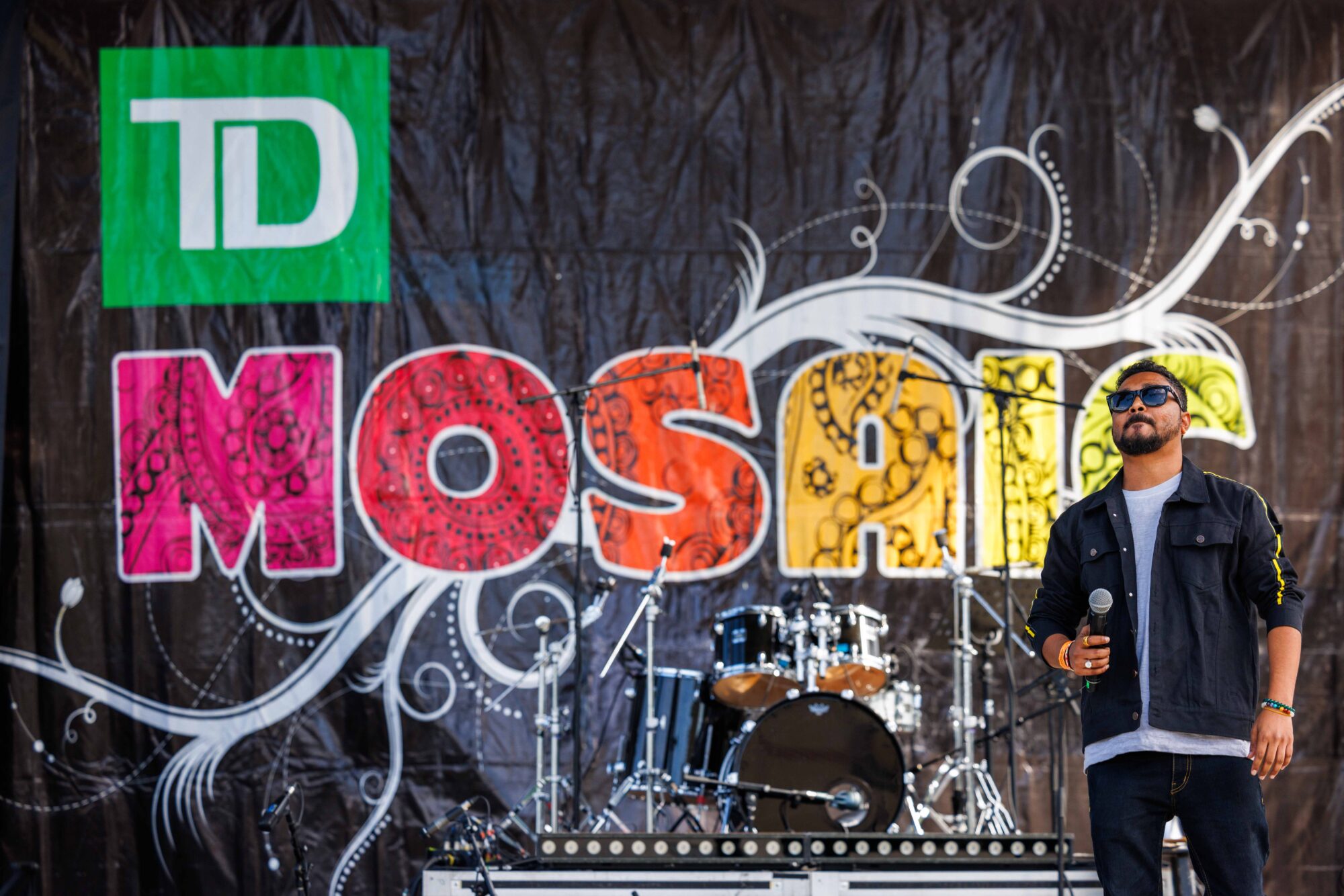 A singer on the TD Mosaic Festival stage at Celebration Square Mississauga, with the patterned MOSAIC backdrop and drum kit behind him.