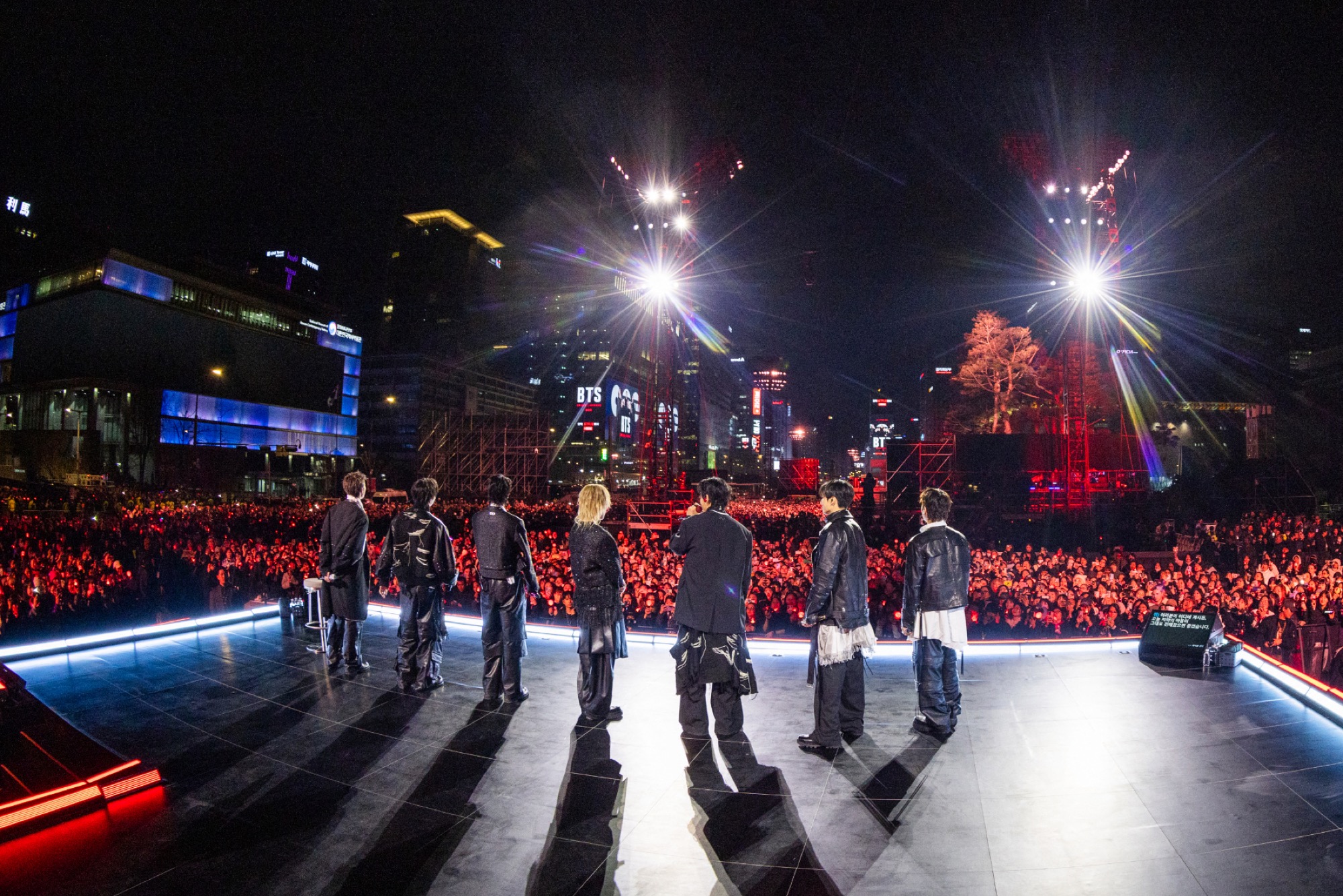BTS members stand on the Gwanghwamun stage looking out over a packed nighttime crowd, with spotlights and city buildings lit for the Netflix comeback live.