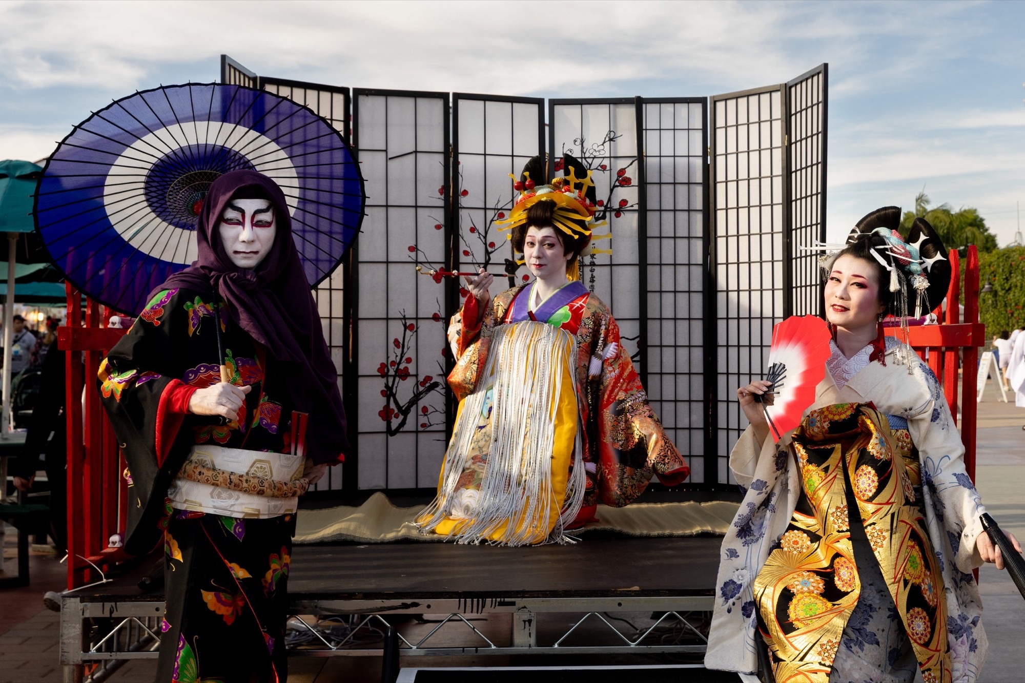 Three performers in elaborate traditional Japanese stage dress pose with a parasol and fan on an outdoor festival stage.