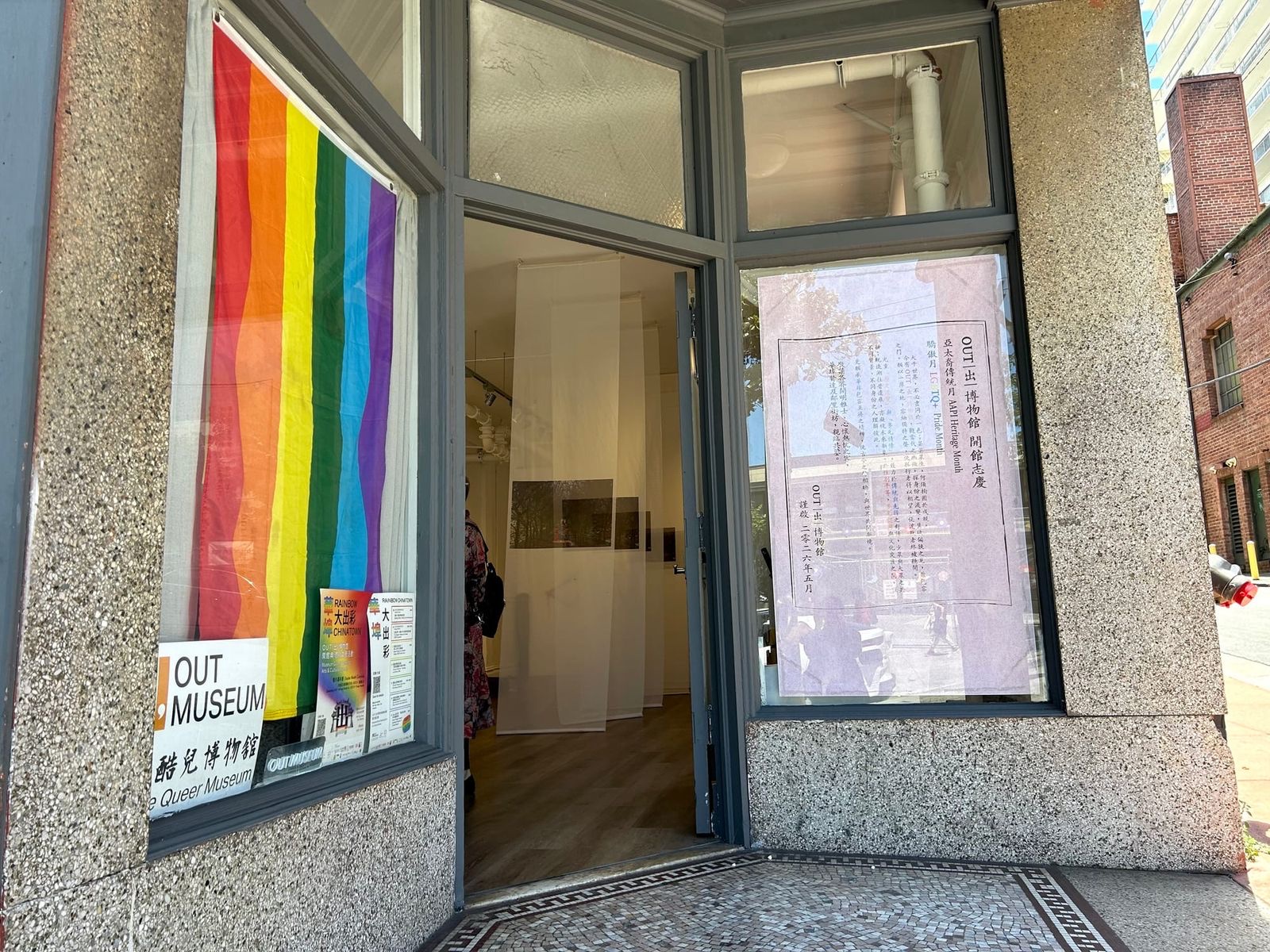 OUT Museum storefront on Clay Street in San Francisco Chinatown, with a rainbow flag in the window, bilingual Queer Museum signage, and hanging exhibition panels visible through the open door.
