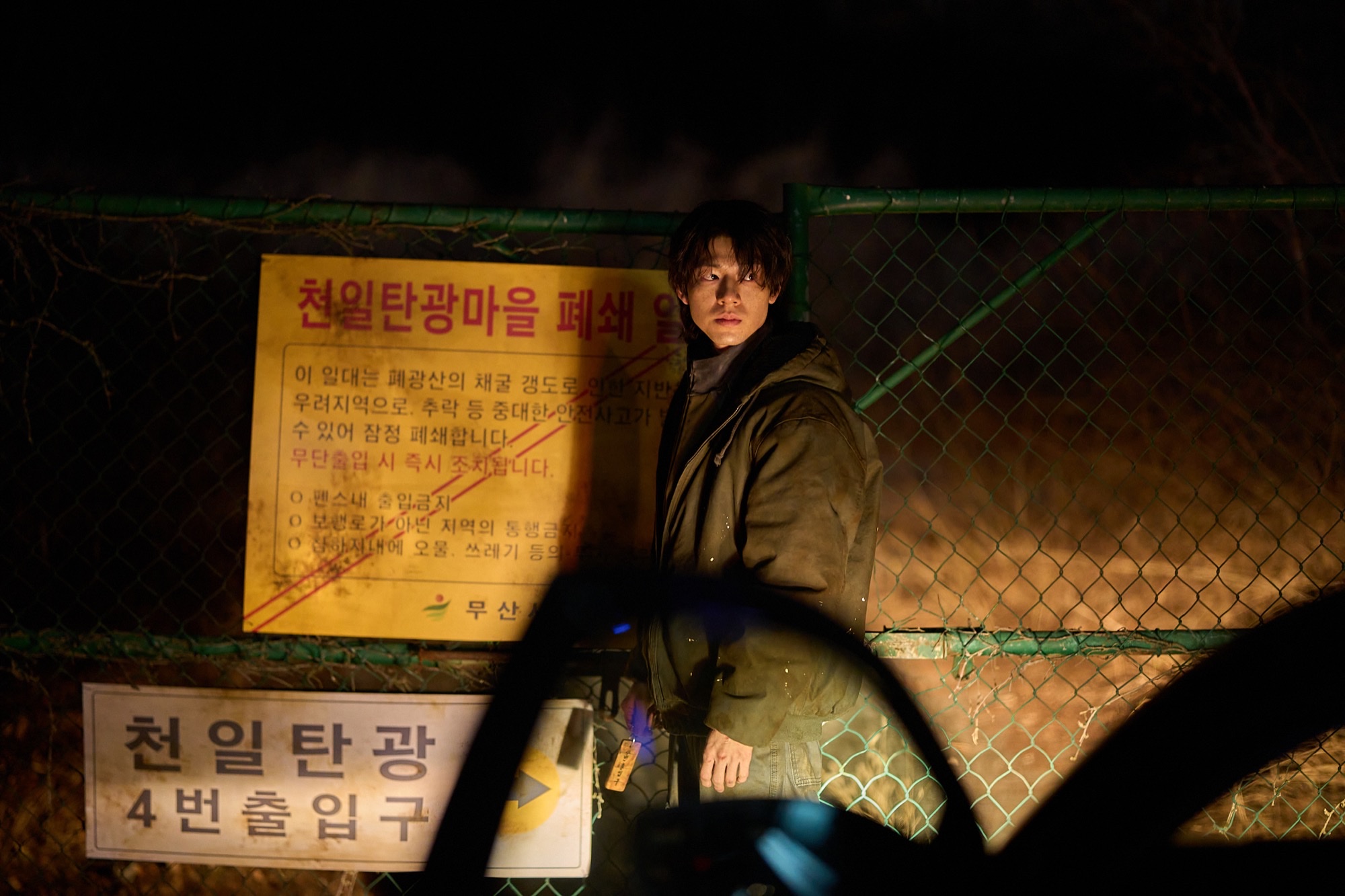 Bae Hyun-sung as Ryu Sun-woo in Paradise Lost, standing at night by a chain-link fence and a yellow mine-closure sign.