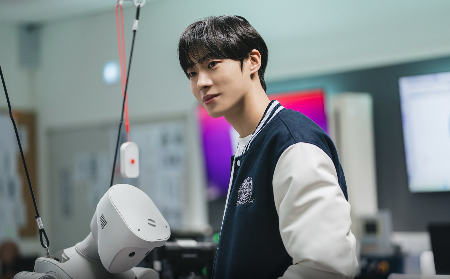 Choo Young-woo as Park Min-jae in a navy varsity jacket in a robotics lab still from Between Steps, with a white research robot in the foreground.