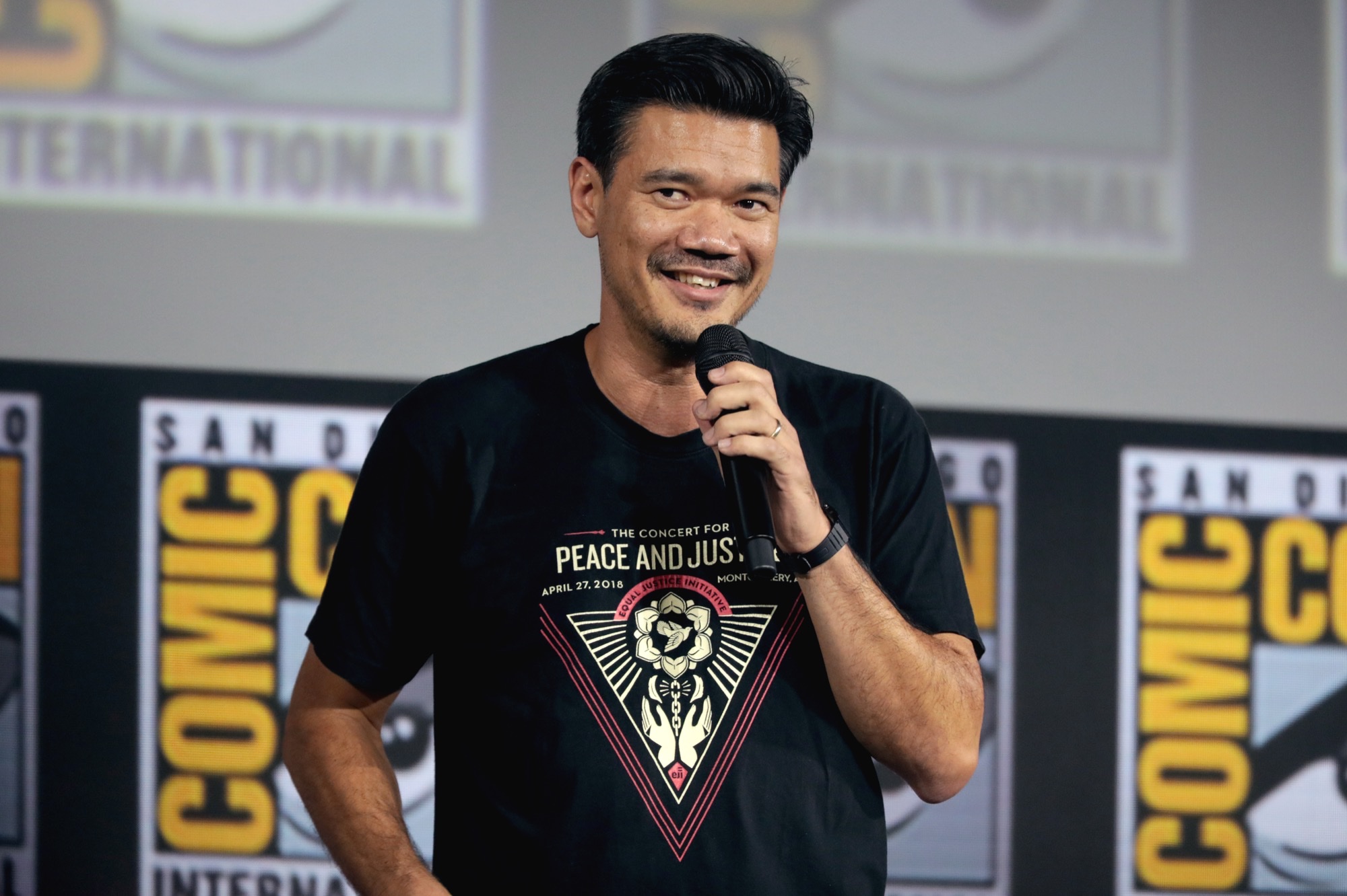 Destin Daniel Cretton smiles while holding a microphone at San Diego Comic-Con, wearing a black Equal Justice Initiative concert shirt.